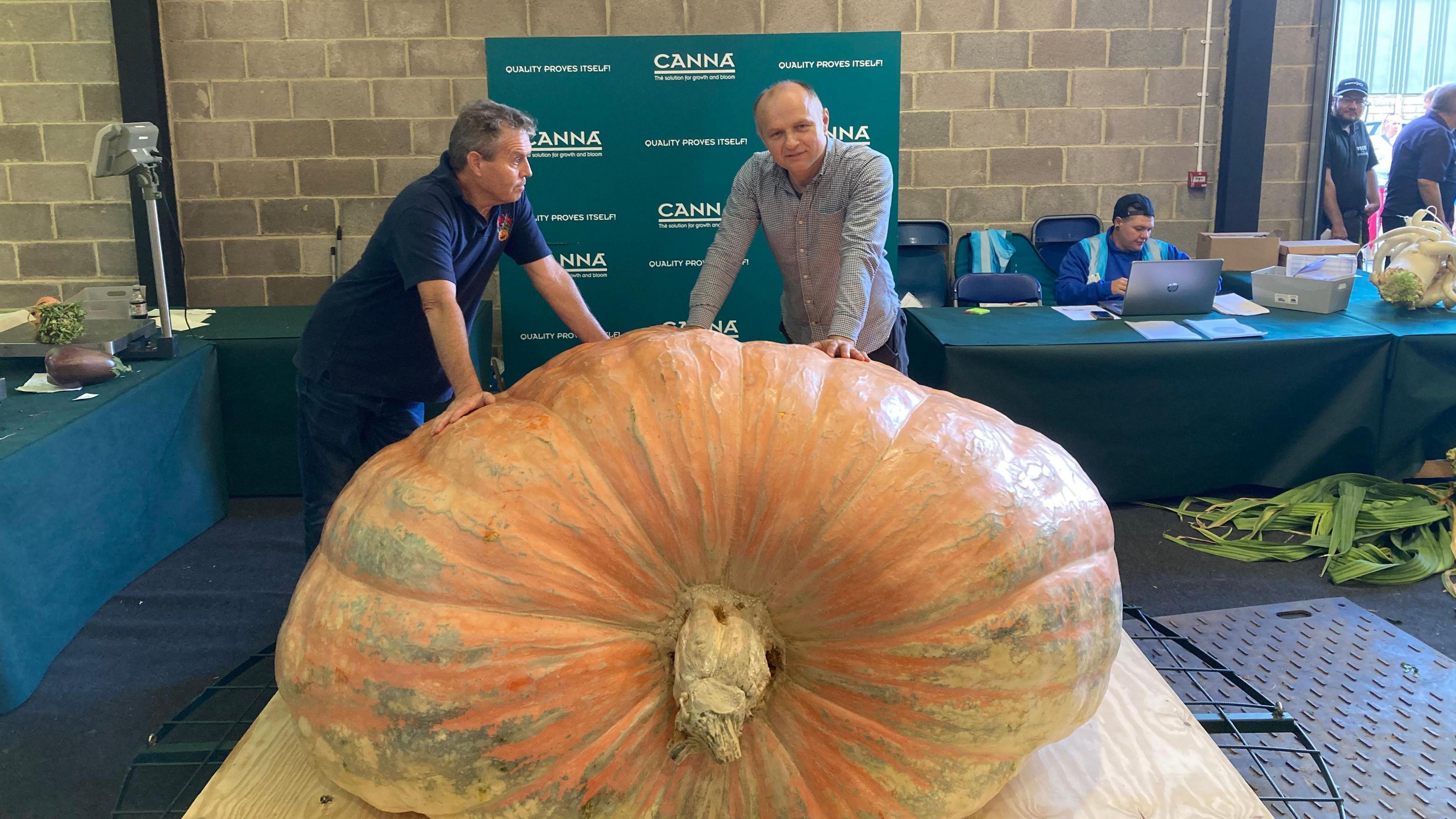 Two men lean against a giant pumpkin which is sat on a wooden board, with another man working at a laptop in the background, sat at a table with a green table cloth over it.