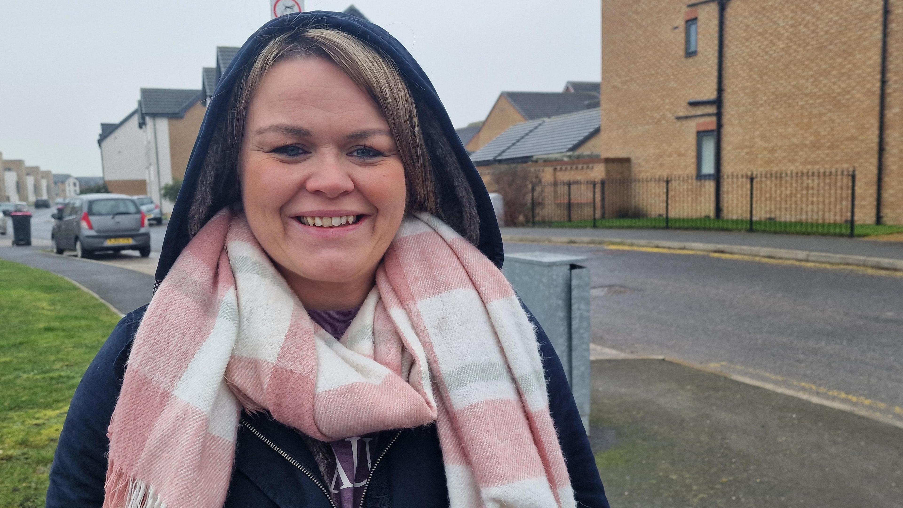 A woman smiles. She is standing on a pavement at the edge of a road on a modern housing development and wears and blue hooded coat and a pink and white checked scarf.