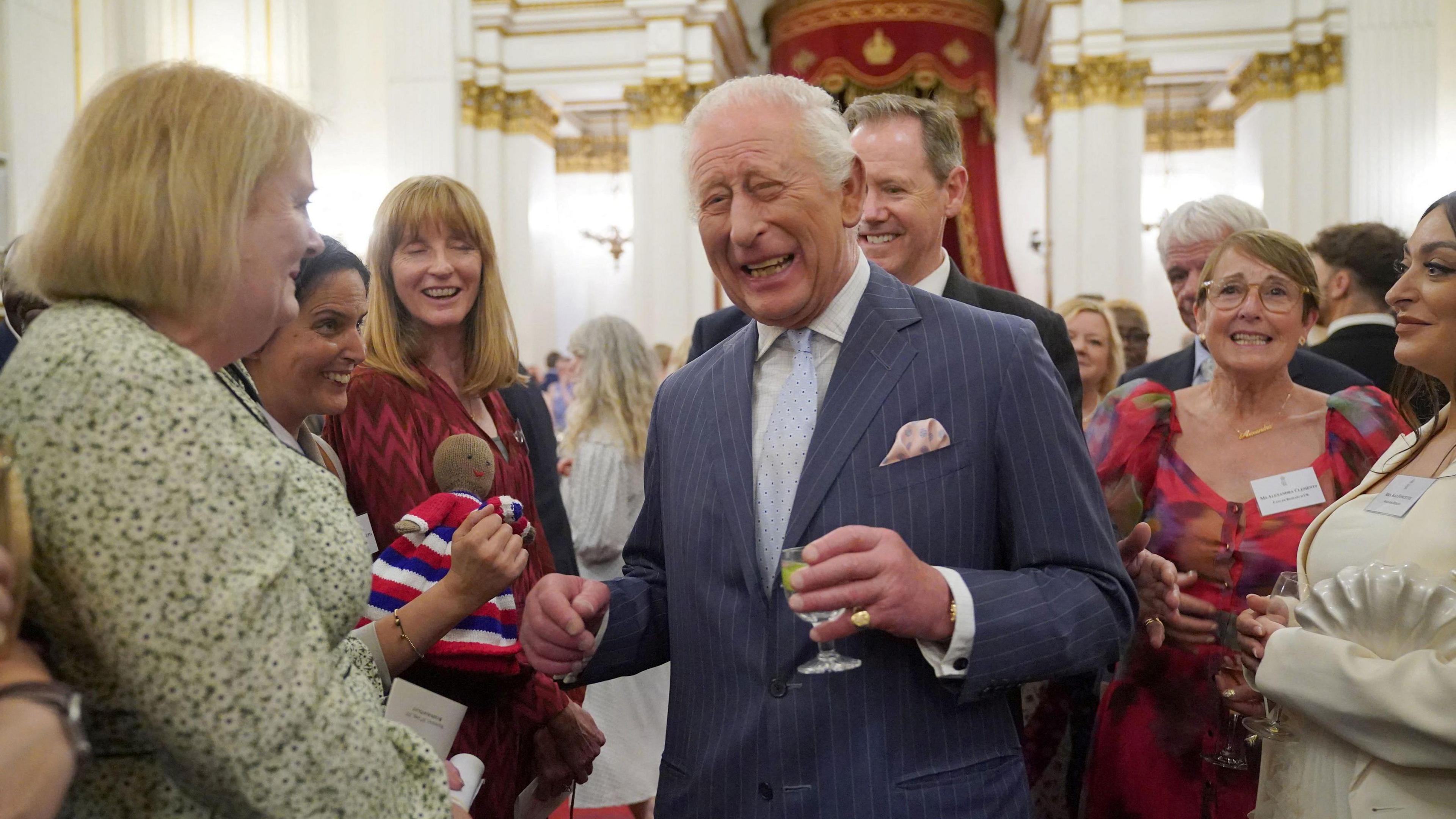 King Charles laughs amongst a smiling group of people at his Buckingham Palace reception for cancer charities. One woman holds up a knitted doll that has no hair.