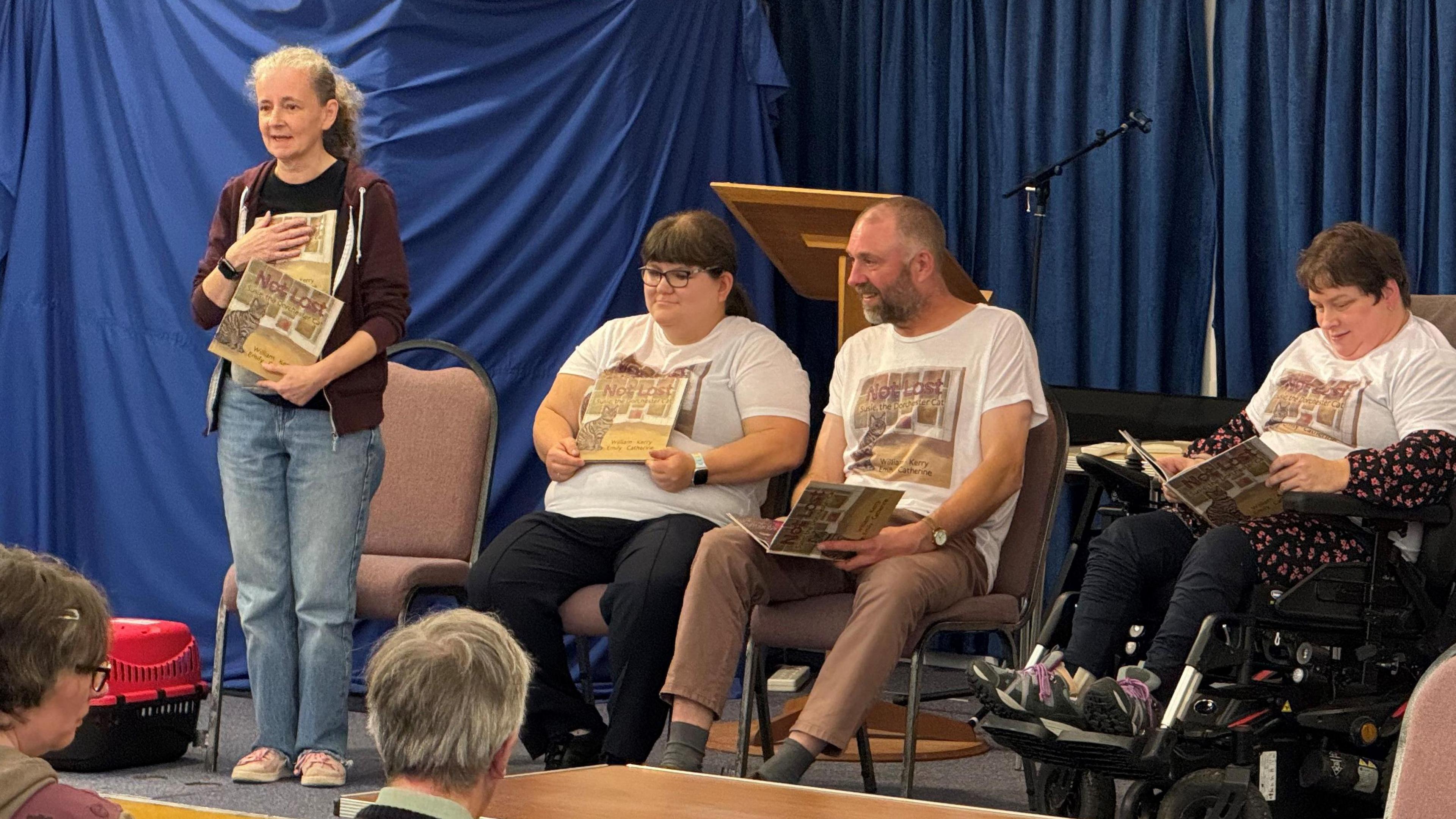 Catherine Owen standing on stage at the book launch. Sitting next to her are Kerry Martin, William Parmiter and Emily Burr. All four of them are holding a copy of the book and wearing a t-shirt bearing an image of the book cover.