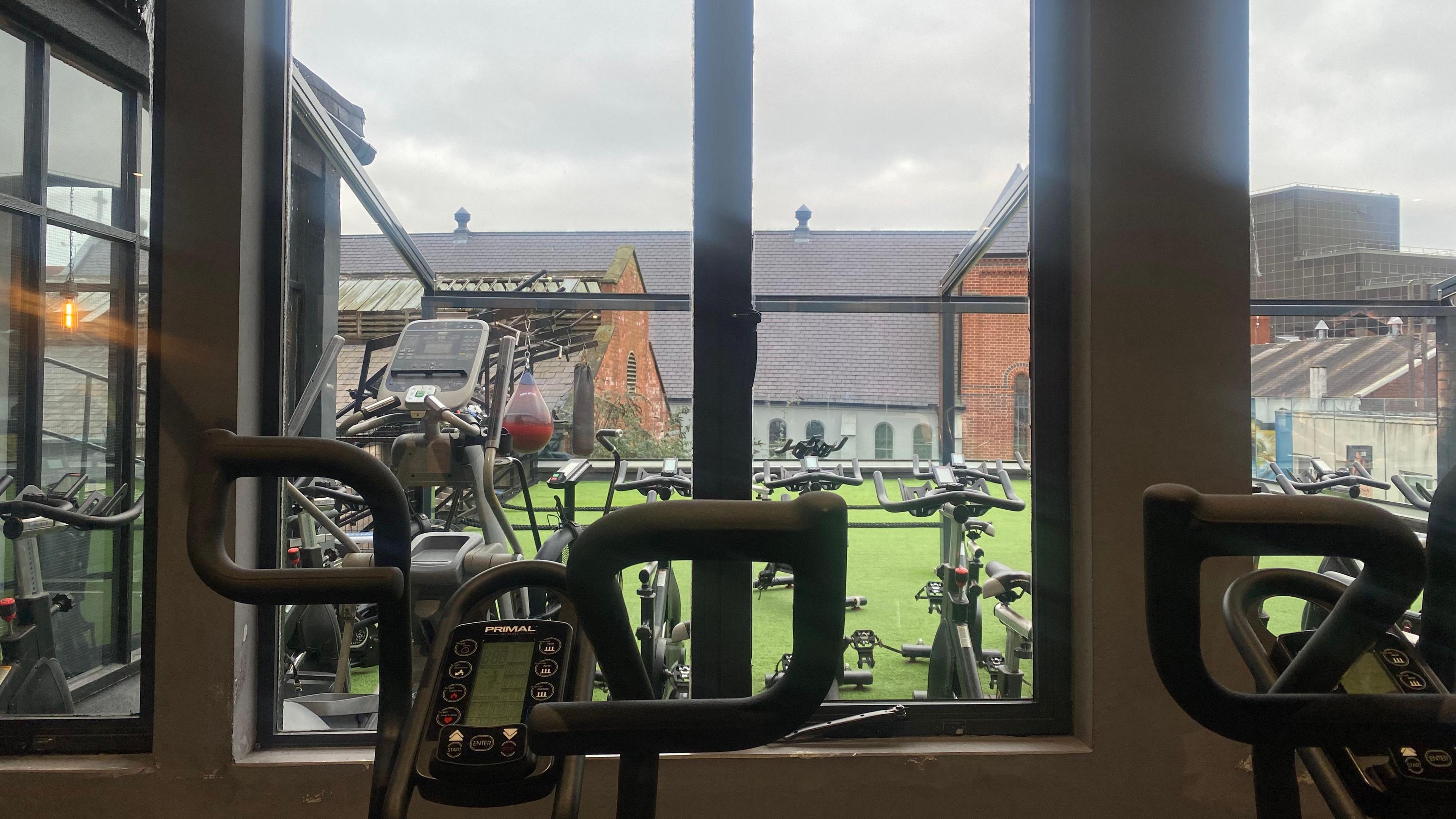 Interior of a gym looking out at a rooftop gym space. There are peleton bikes inside and outside. Outside there is artificial grass and a view of city rooftops.