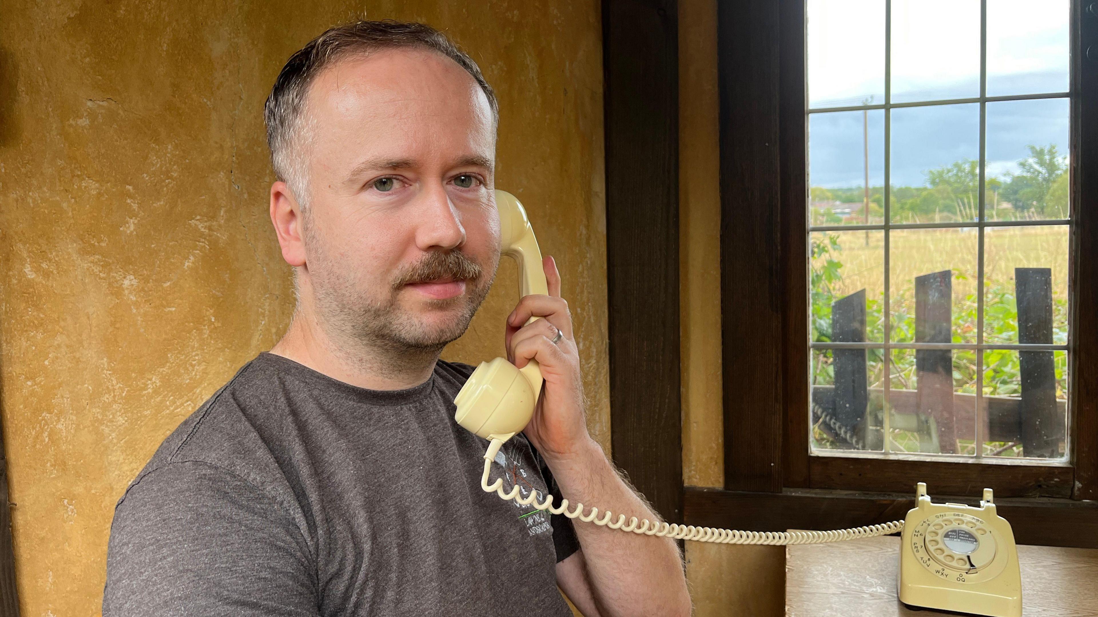 A man with short hair and a moustache sits indoors holding the receiver of a cream rotary telephone, with the phone resting on a wooden table beside him.