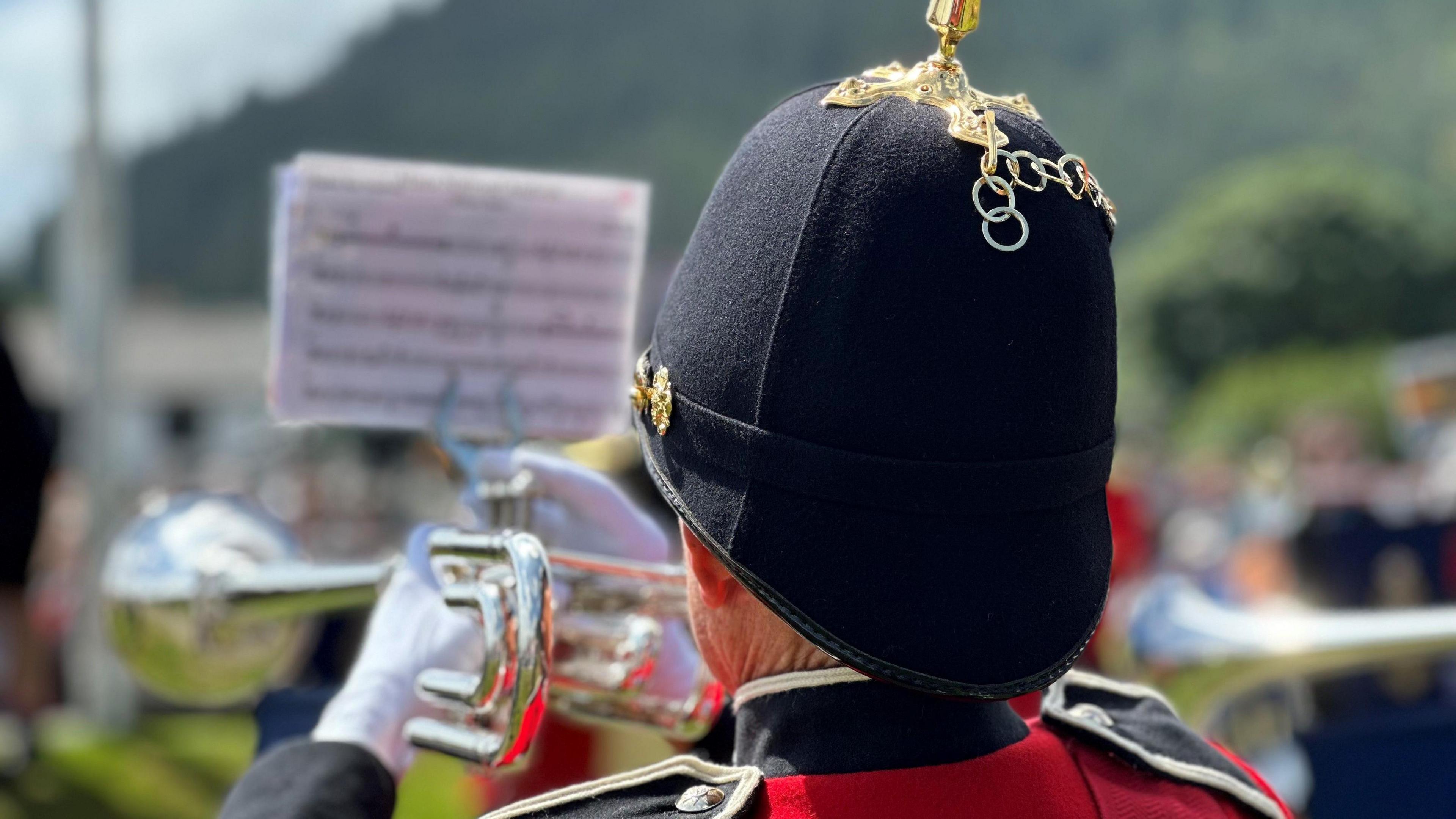 A trumpeter from the back. He is wearing a black helmet featuring a gold spike and white gloves. There is a music stand holding music attached to the trumpet.