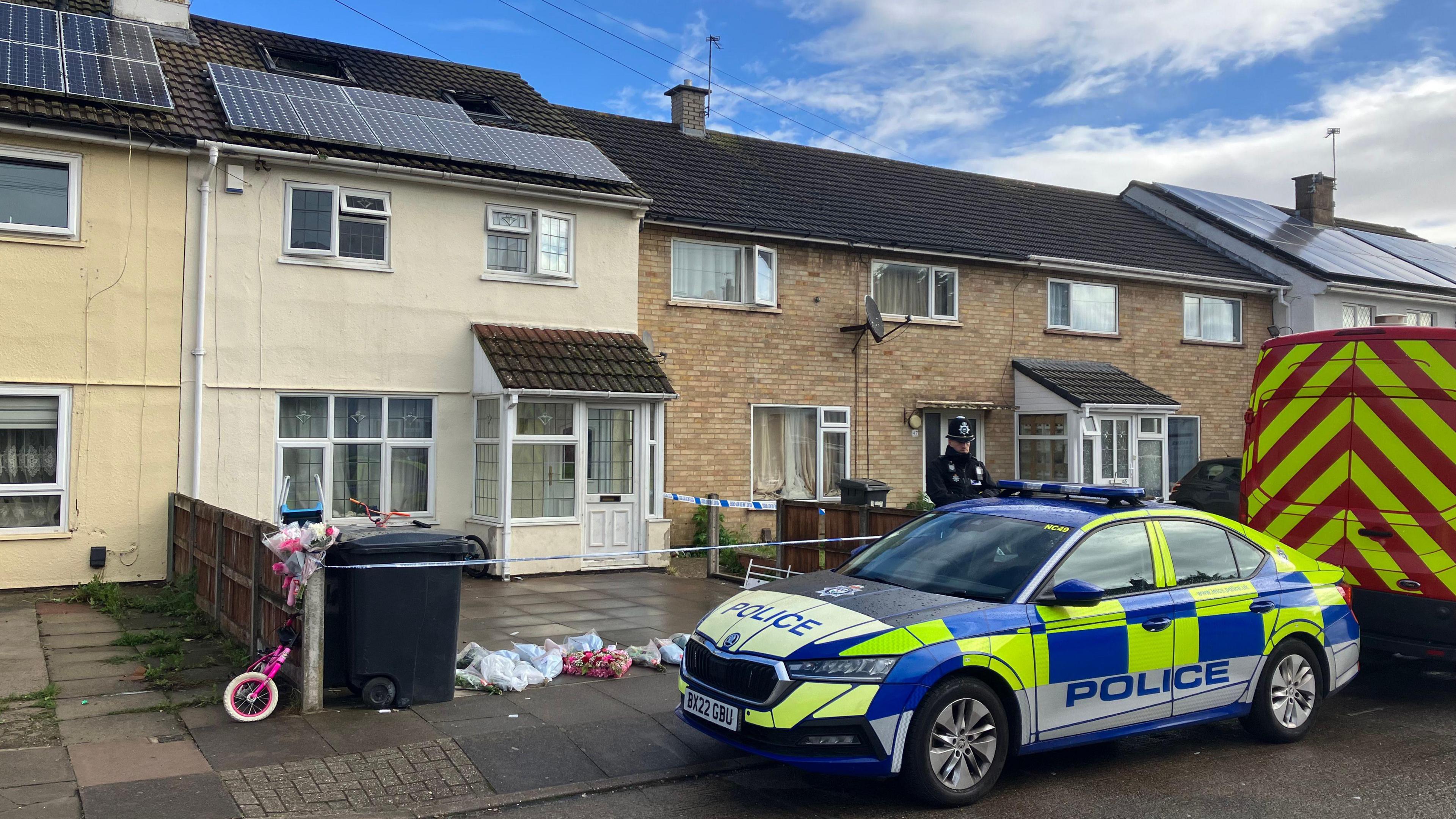 Terraced house surrounded by police tape, with an officer and police car outside. A pink child's bike and flowers can be seen in front of the house 