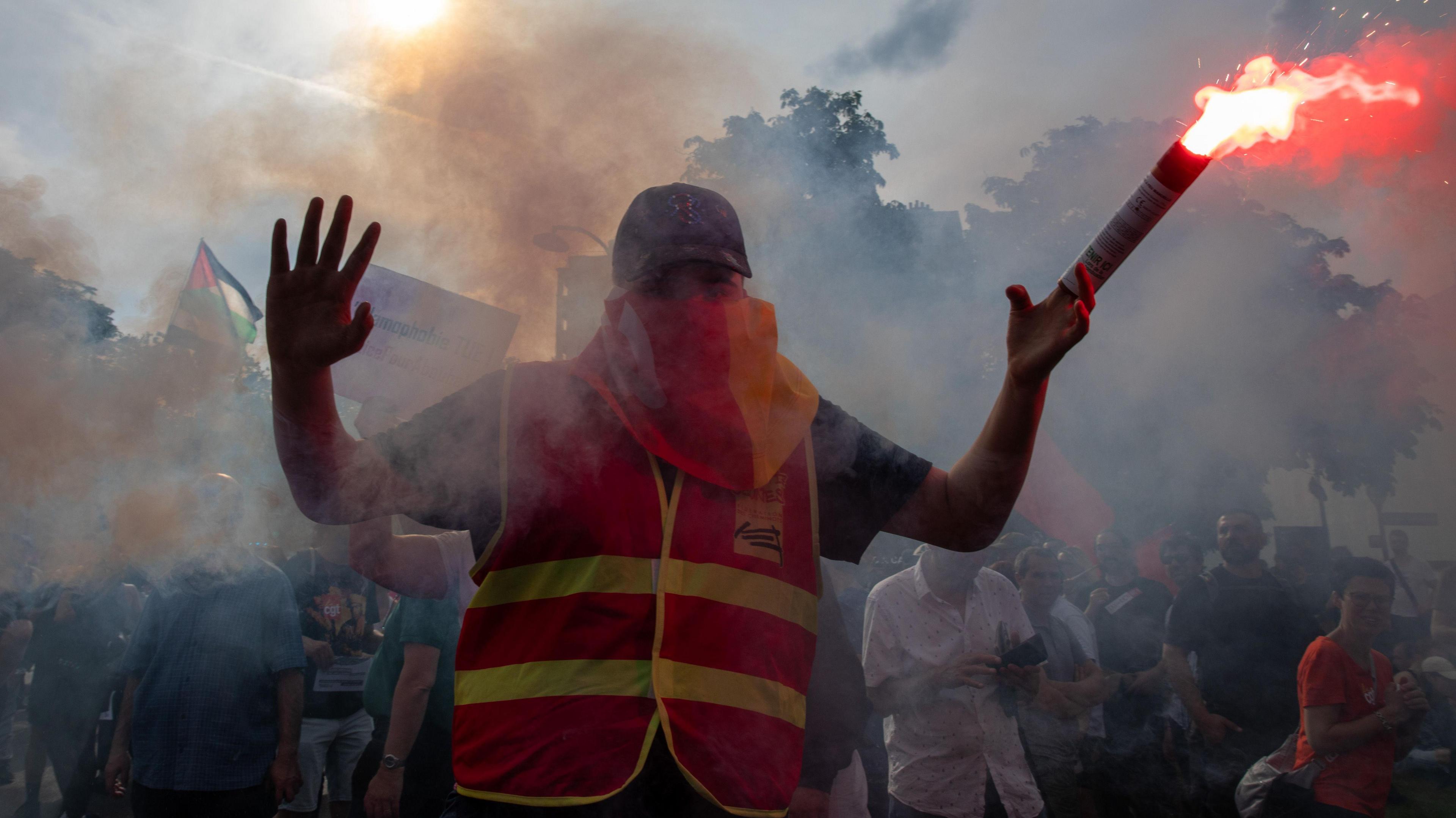 A man in a hi-vis jacket and sunglasses waves a lit, red flare at a protest in France