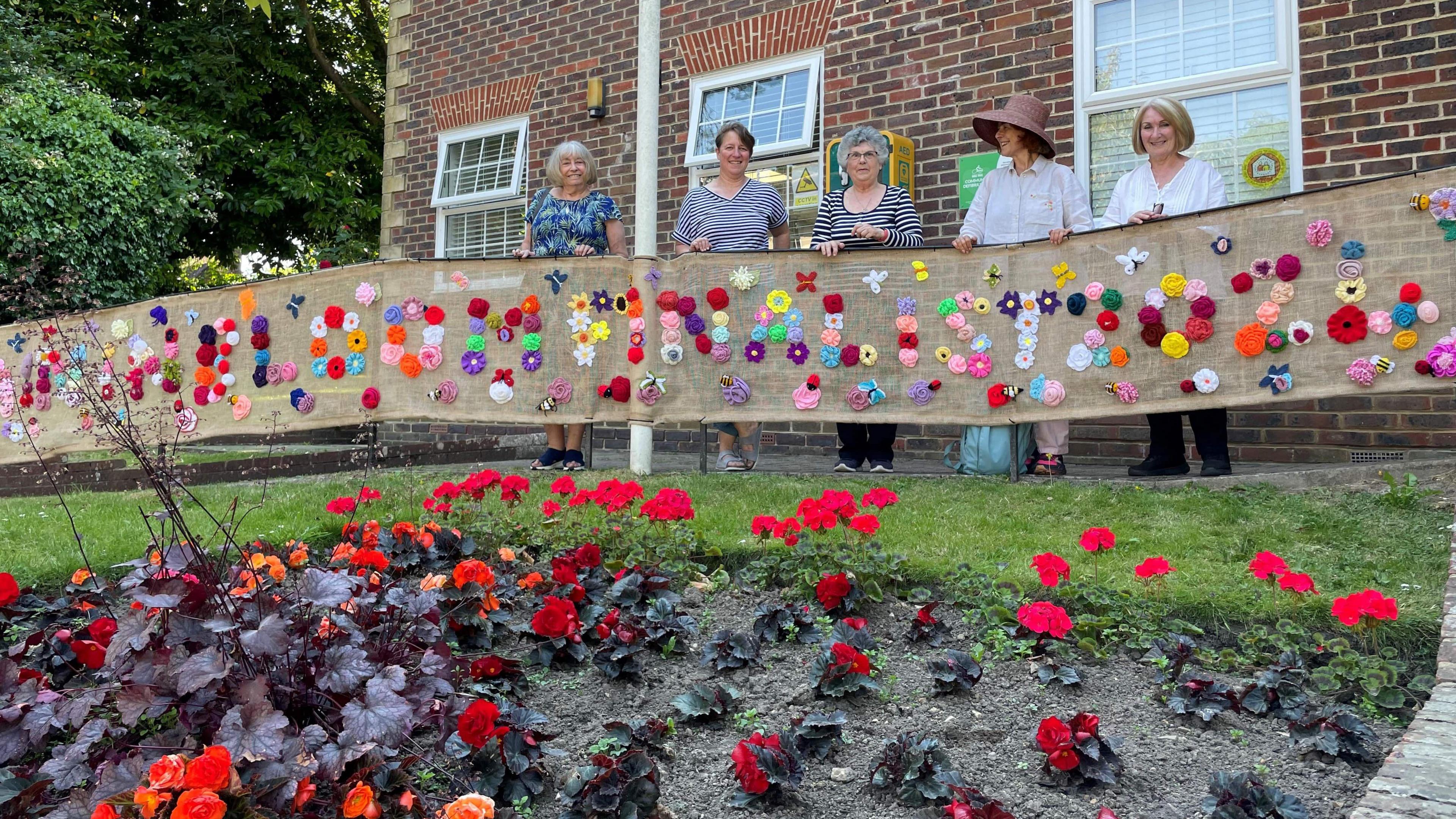 Five women stand behind a flowely banner behind a flower bed