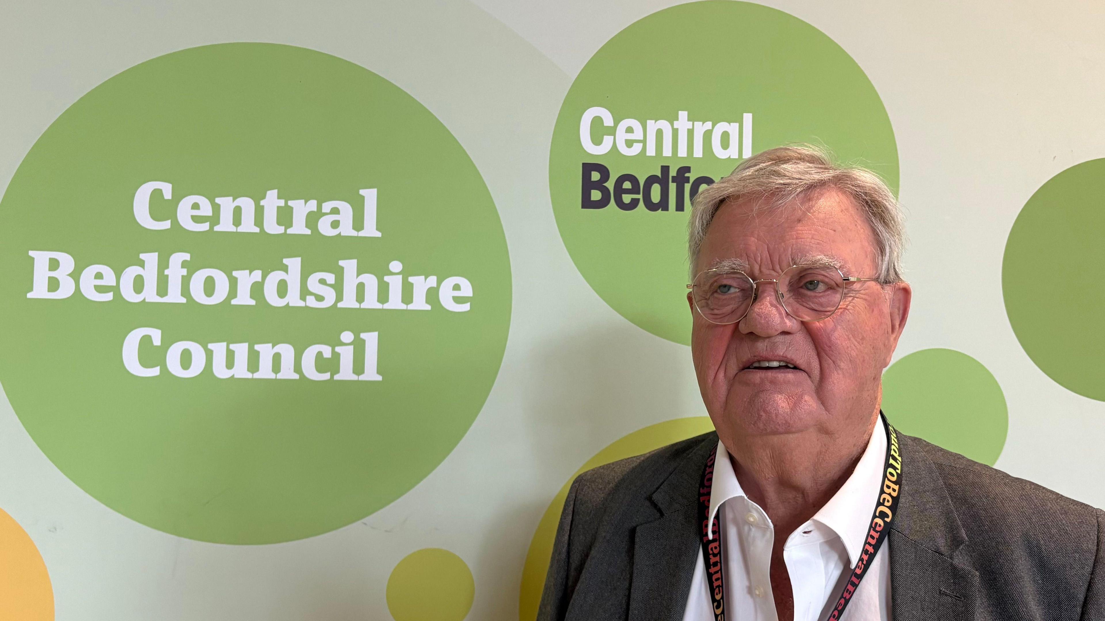 Steve Owen with short, light brown hair, wearing a brown jacket, white shirt and red lanyard, standing in front of a board showing the green Central Bedfordshire Council logo
