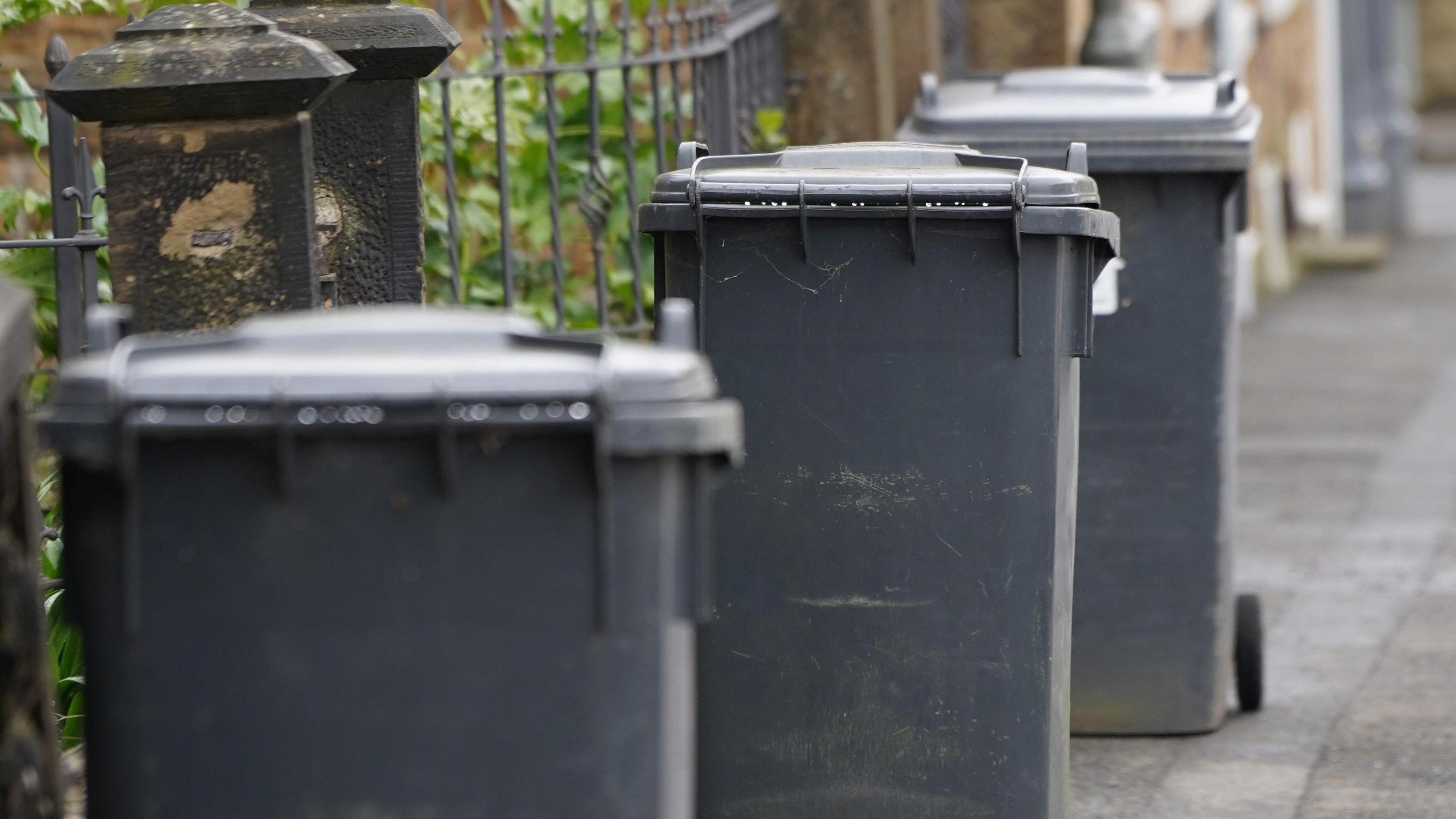 Three regular-sized grey wheelie bins on the pavement outside some gates.