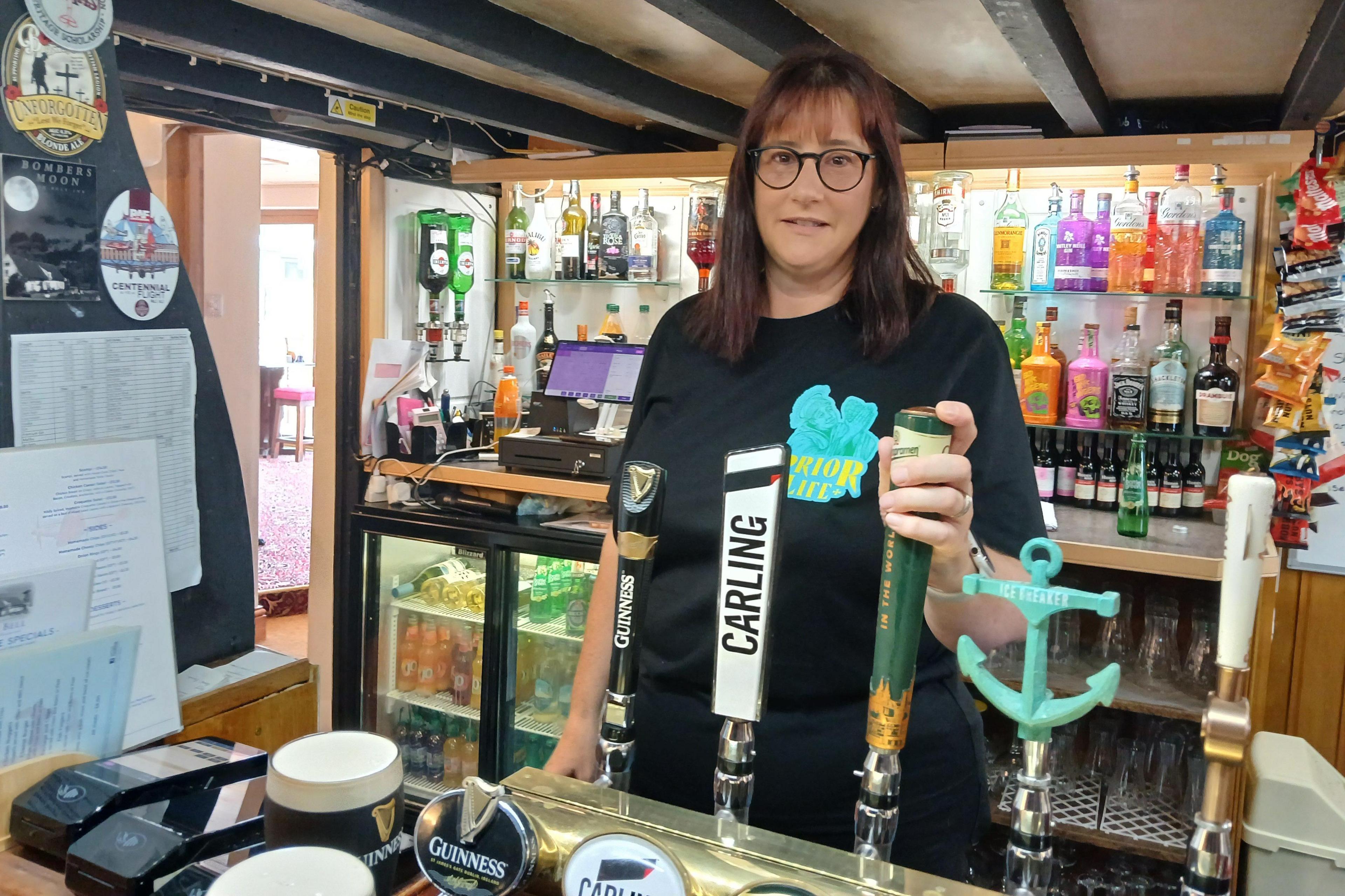 Sue Maltby stands behind the bar of the Blue Bell Inn. She has shoulder-length, dark brown hair and wears a black T-shirt. She rests a hand on one of five beer pumps. In the background are drinks fridges and rows of spirit bottles.