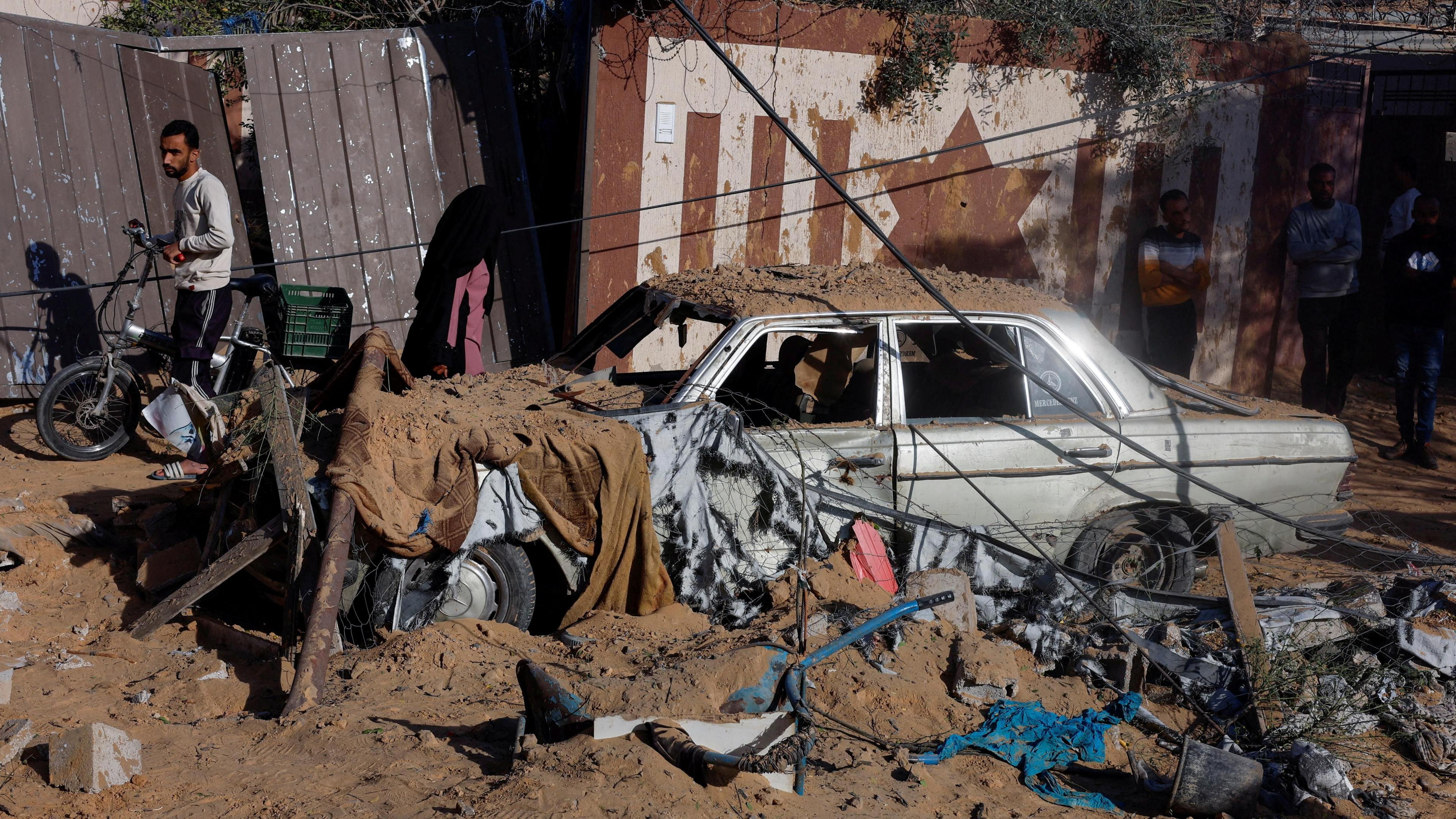 Palestinians stand near a destroyed car at the site of an Israeli strike on a house in Khan Younis, in the southern Gaza Strip (25 October 2024)