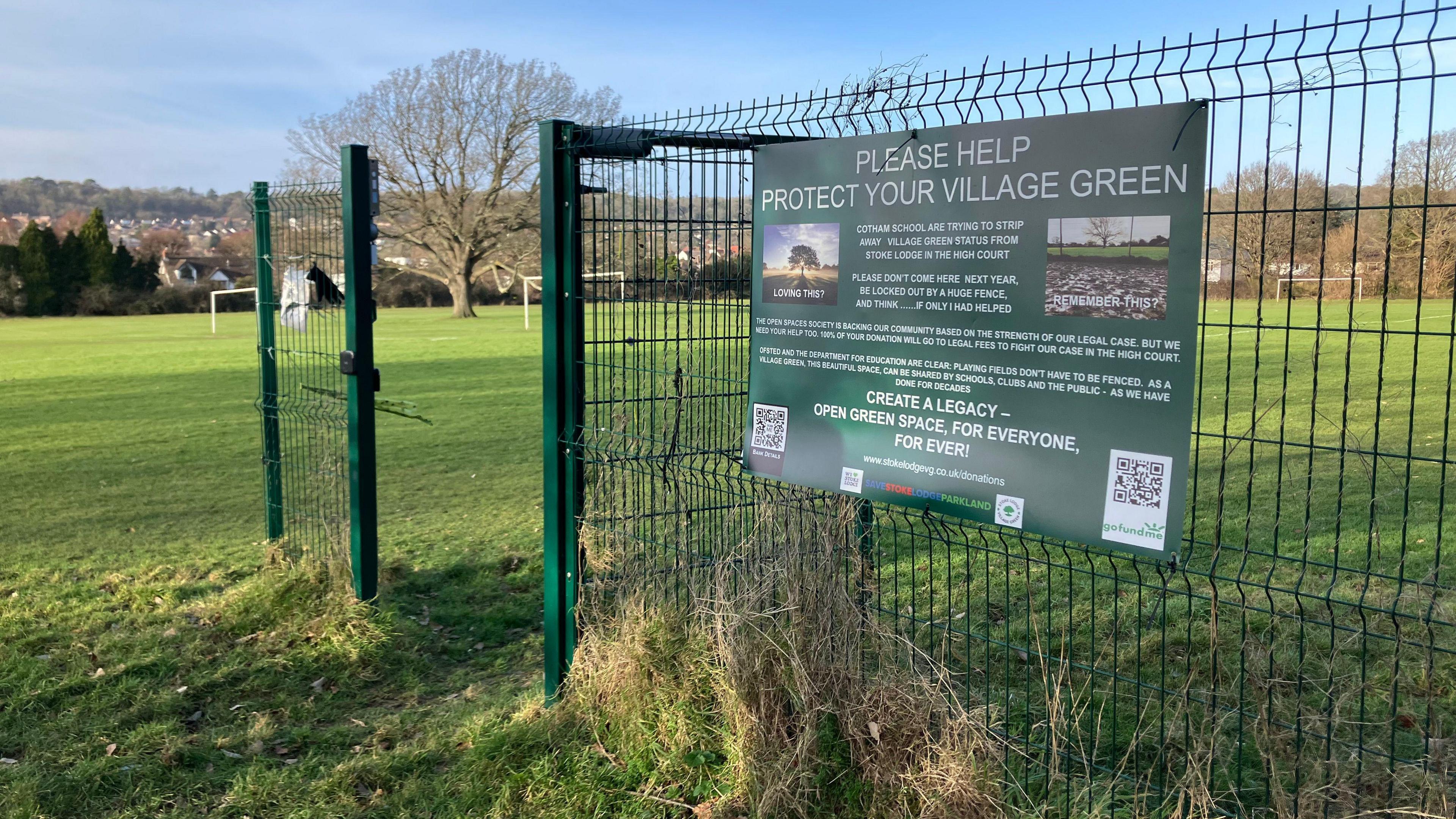 A dark green fence cordons off green space in a playing field with goal posts and trees.