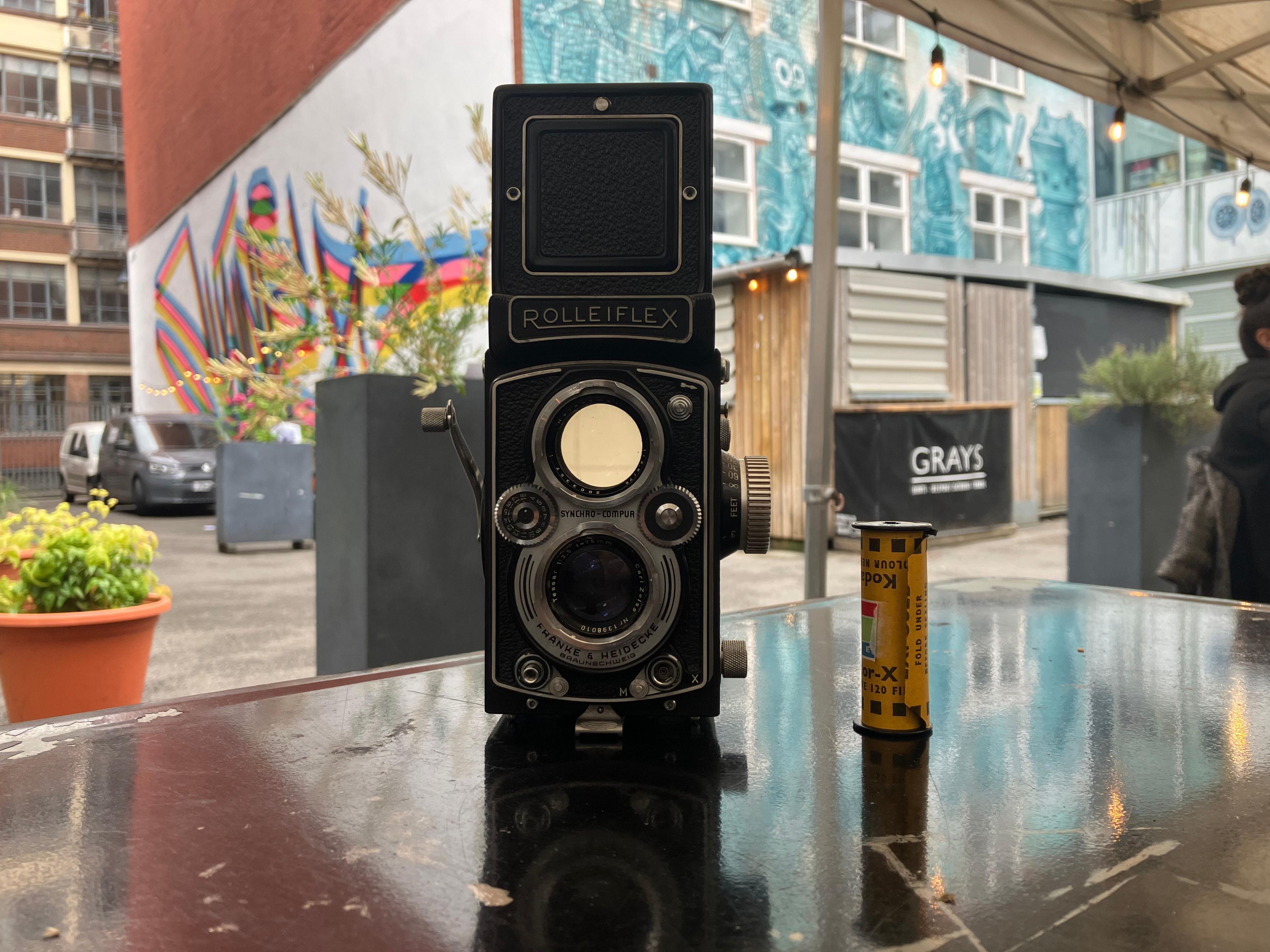 A Rolleiflex camera and a reel of film sit on a table. There is a colourful mural in the background.