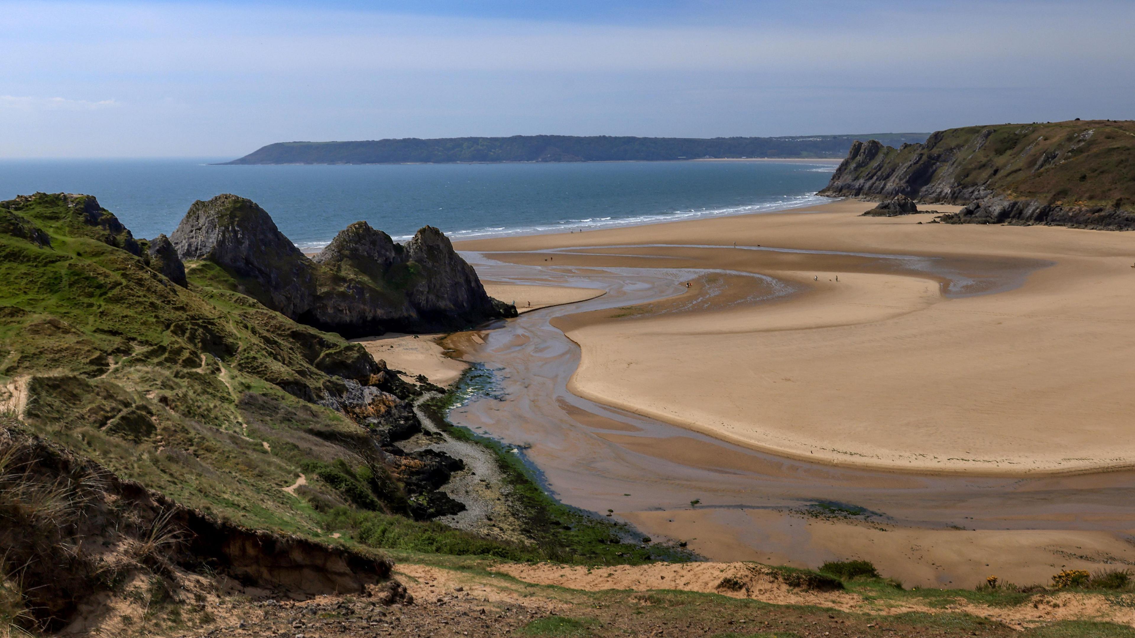 A wide beach with golden sand on a sunny day. The cliffs can be seen in the left and right of the photo, with sand in-between. The blue sea is in the distance, with the coastline of opposite land still visible. 