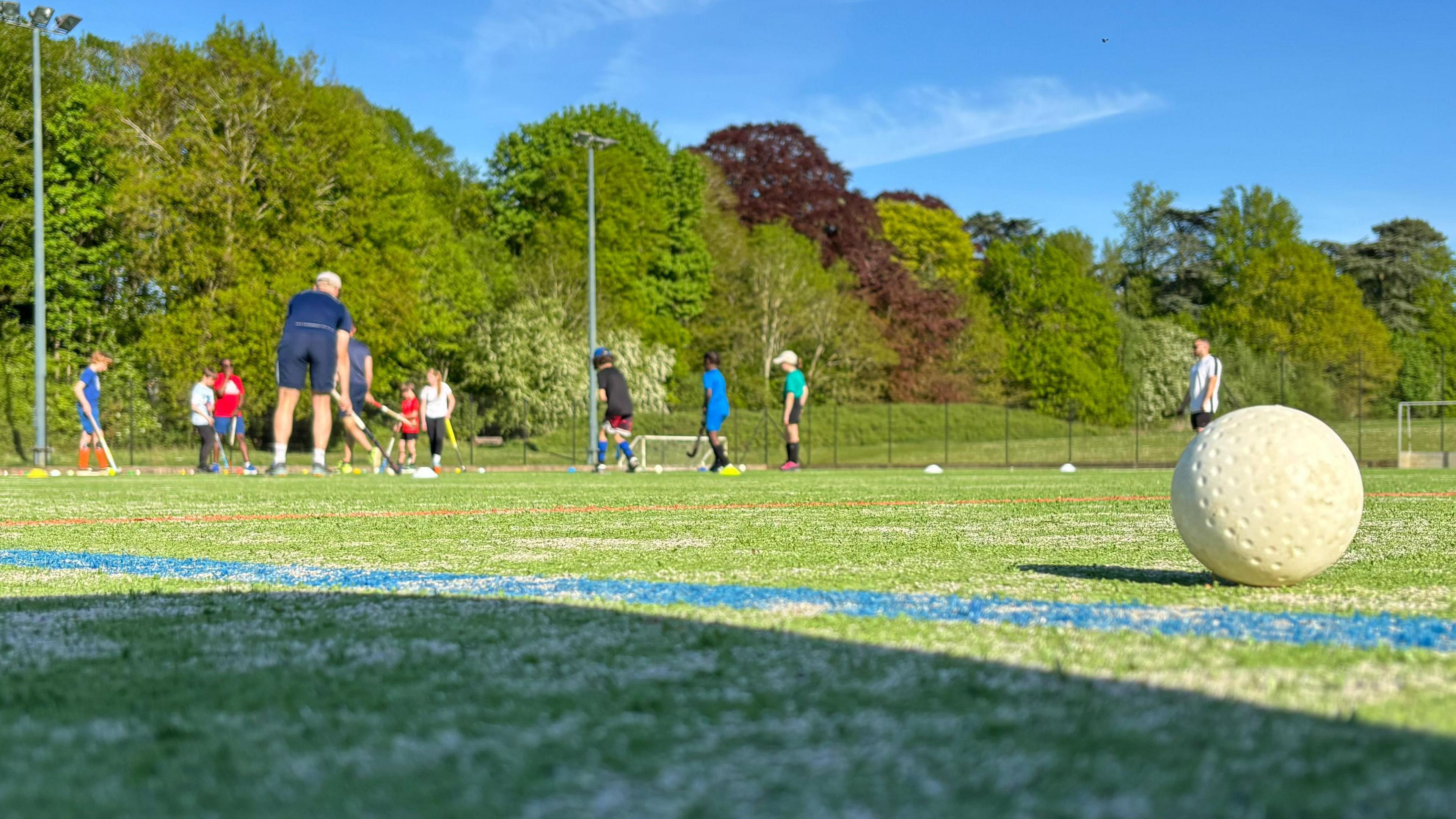 A close-up shot of a white hockey ball on a green pitch with a group of coaches and children playing hockey in the background. The group are dressed in various sports attire.