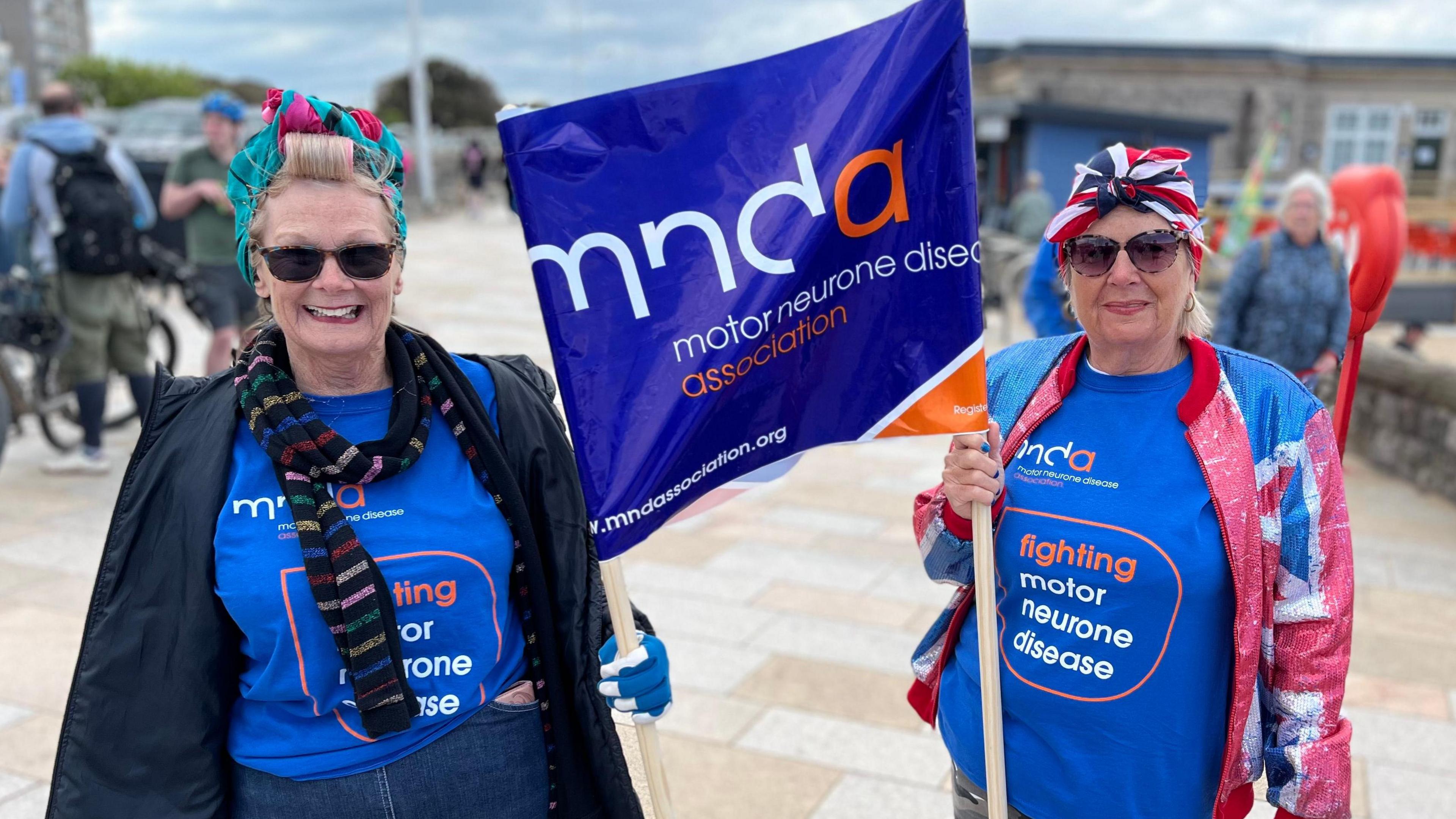 Two women at the tank push event in Weston-super-Mare. They are both carrying a flag and wearing t-shirts with the Motor Neurone Disease Association logo on it.