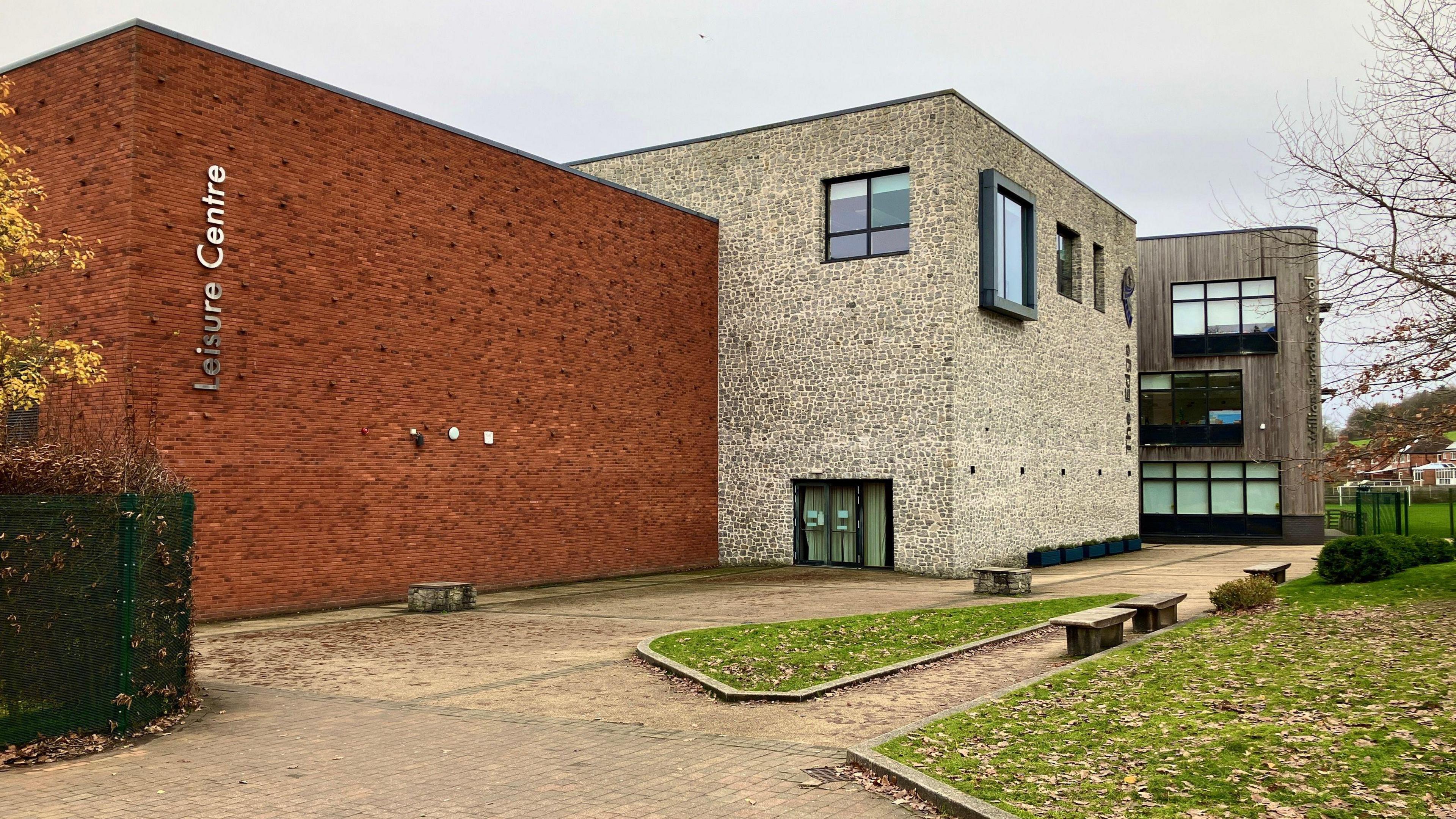 A school with grey stone exterior and adjoining leisure centre, built with red bricks and vertical metal signage reading 'leisure centre'.