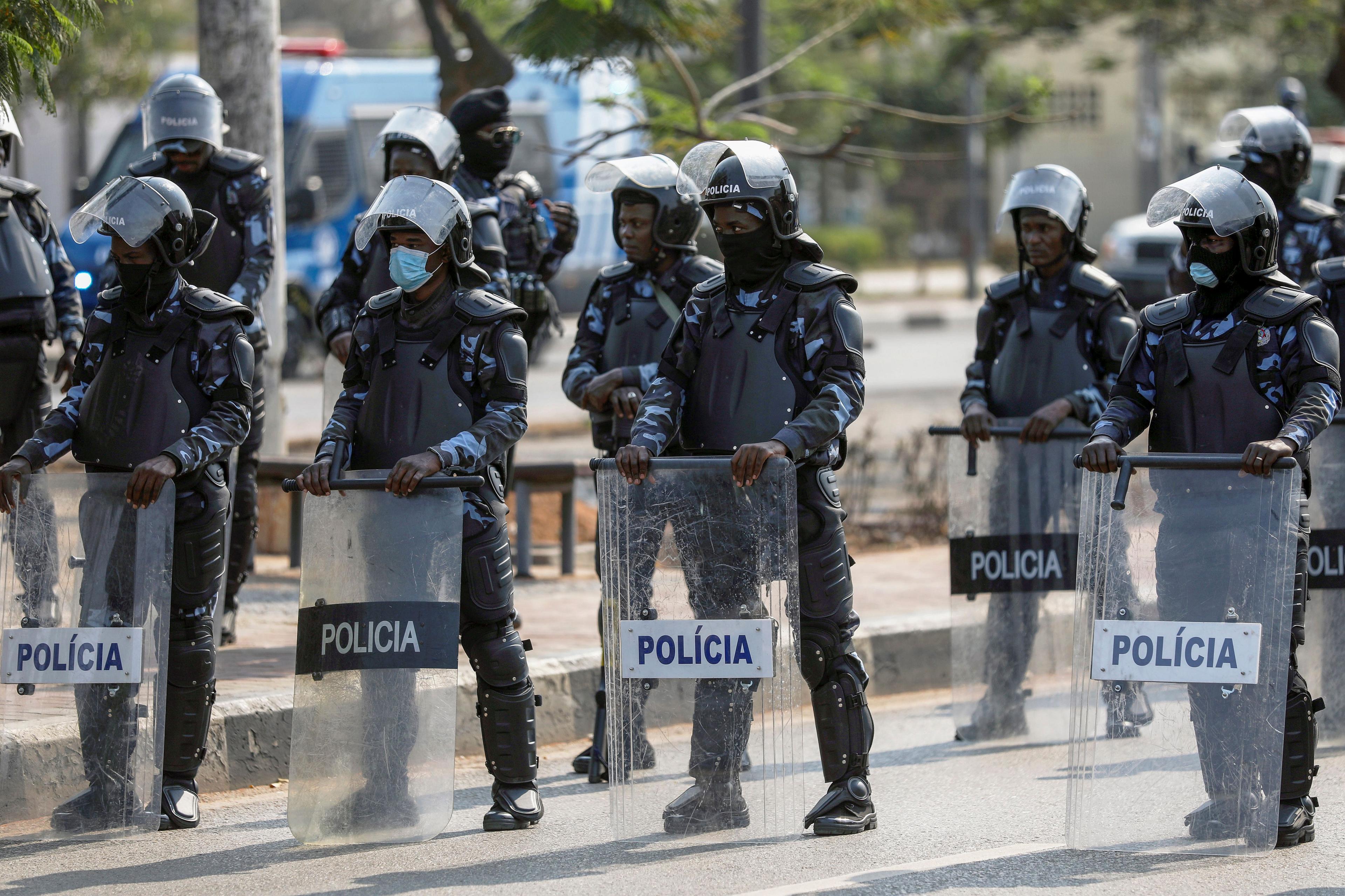Police officers wearing riot gear and shield form a blockade on a road.