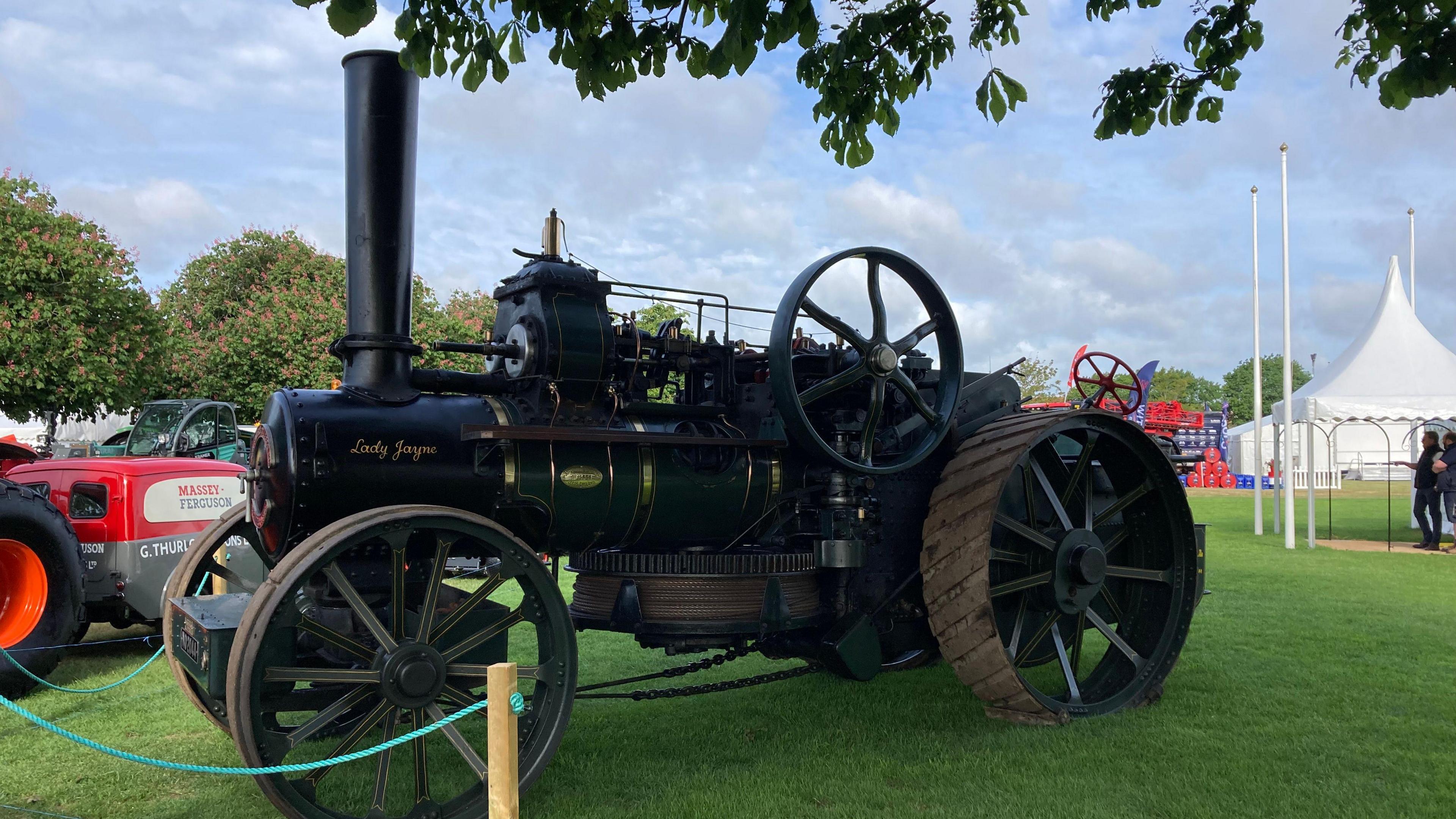 A vintage ploughing vehicle parked in a grass field. It is black and has large wheels as well as a large vertical pipe from its engine on the front.