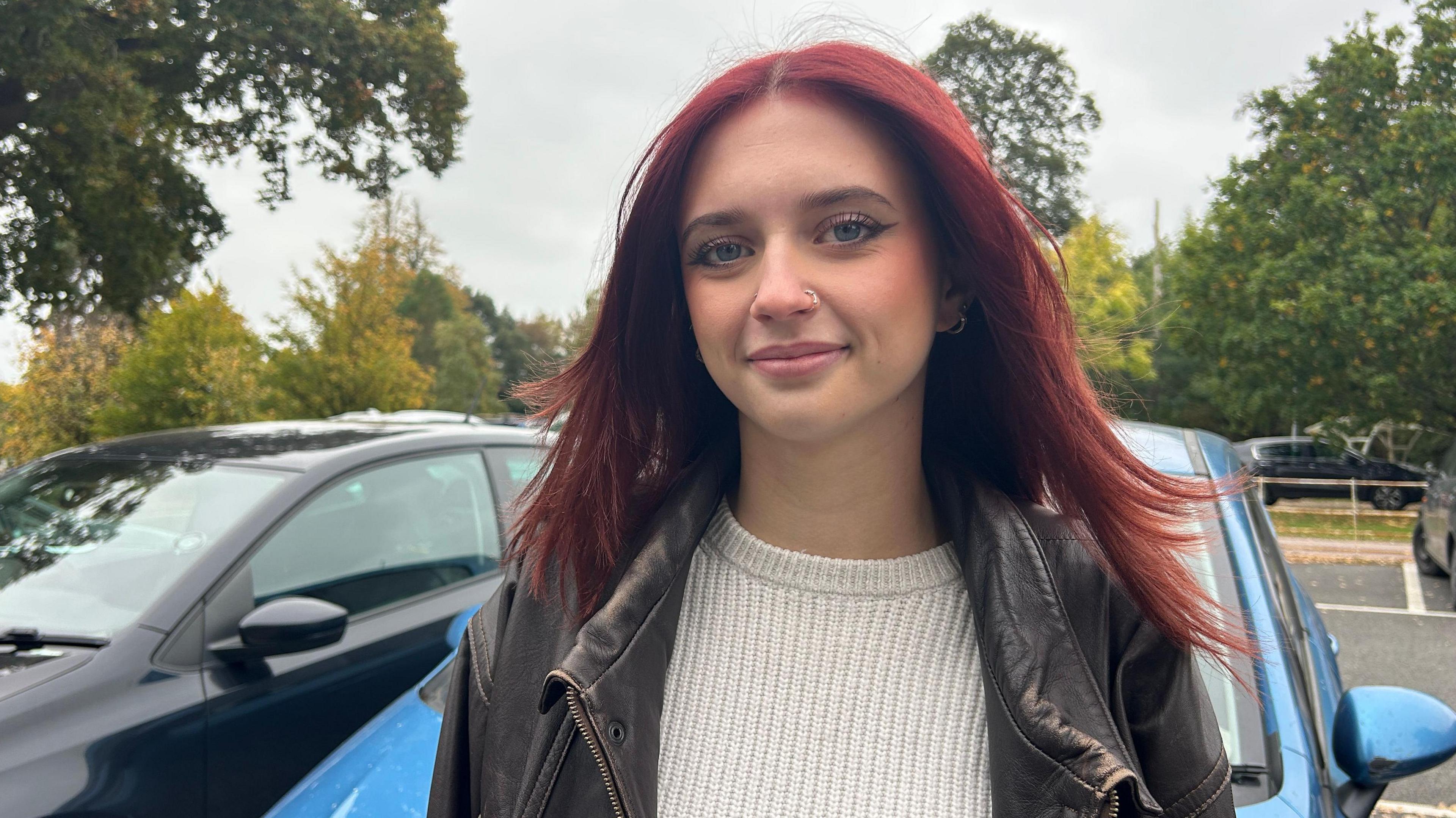 A young woman with long red hair, wearing a cream jumper and a brown leather jacket standing in front of a blue car and smiling.