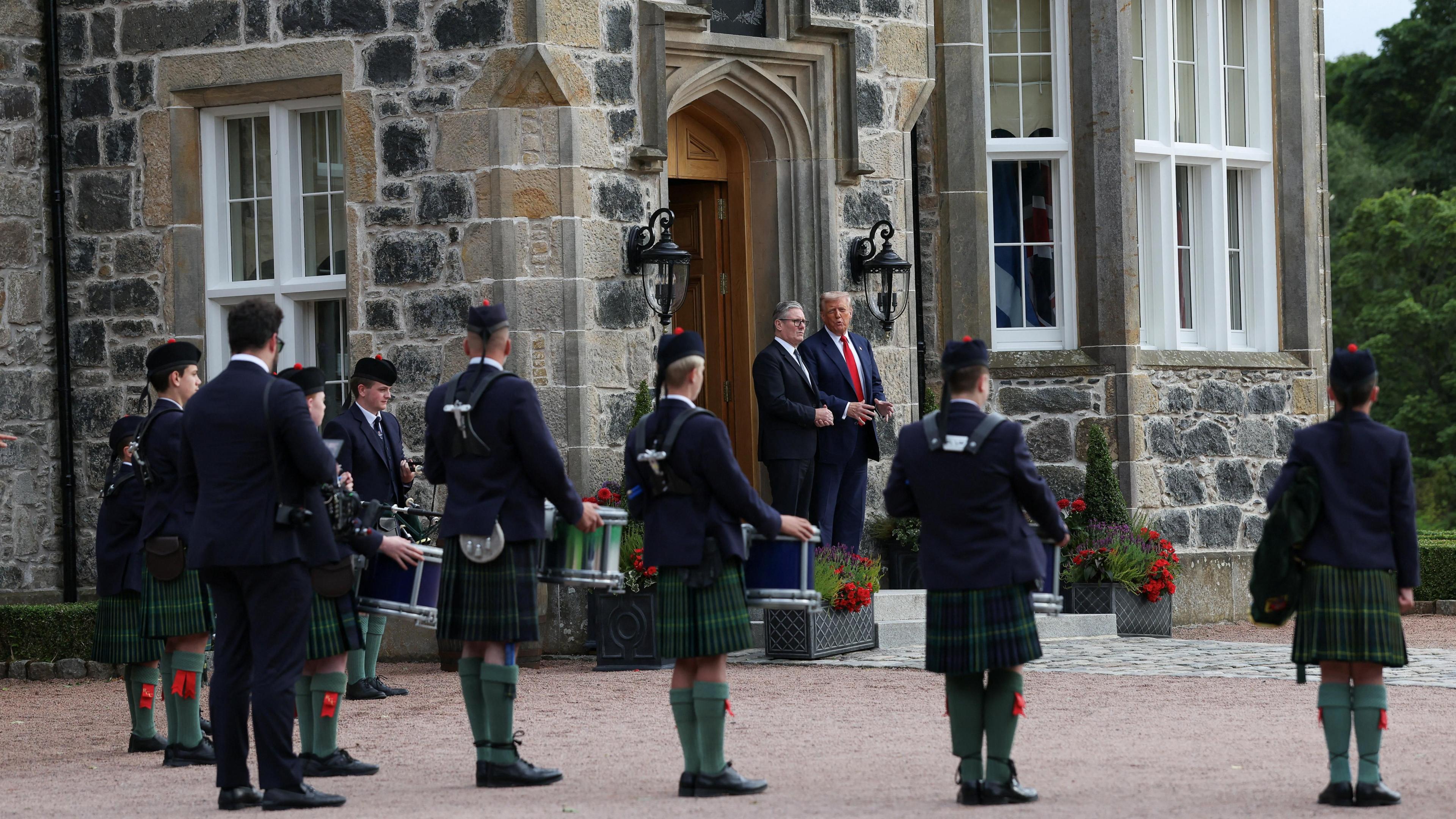 Donald Trump and Keir Starmer are welcomed to McLeod House at the Menie golf resort by the Robert Gordon College pipe band. They are standing in the doorway of the stone building wearing dark suits and the pipers and drummers, wearing traditional Highland dress, are lined up in the foreground.