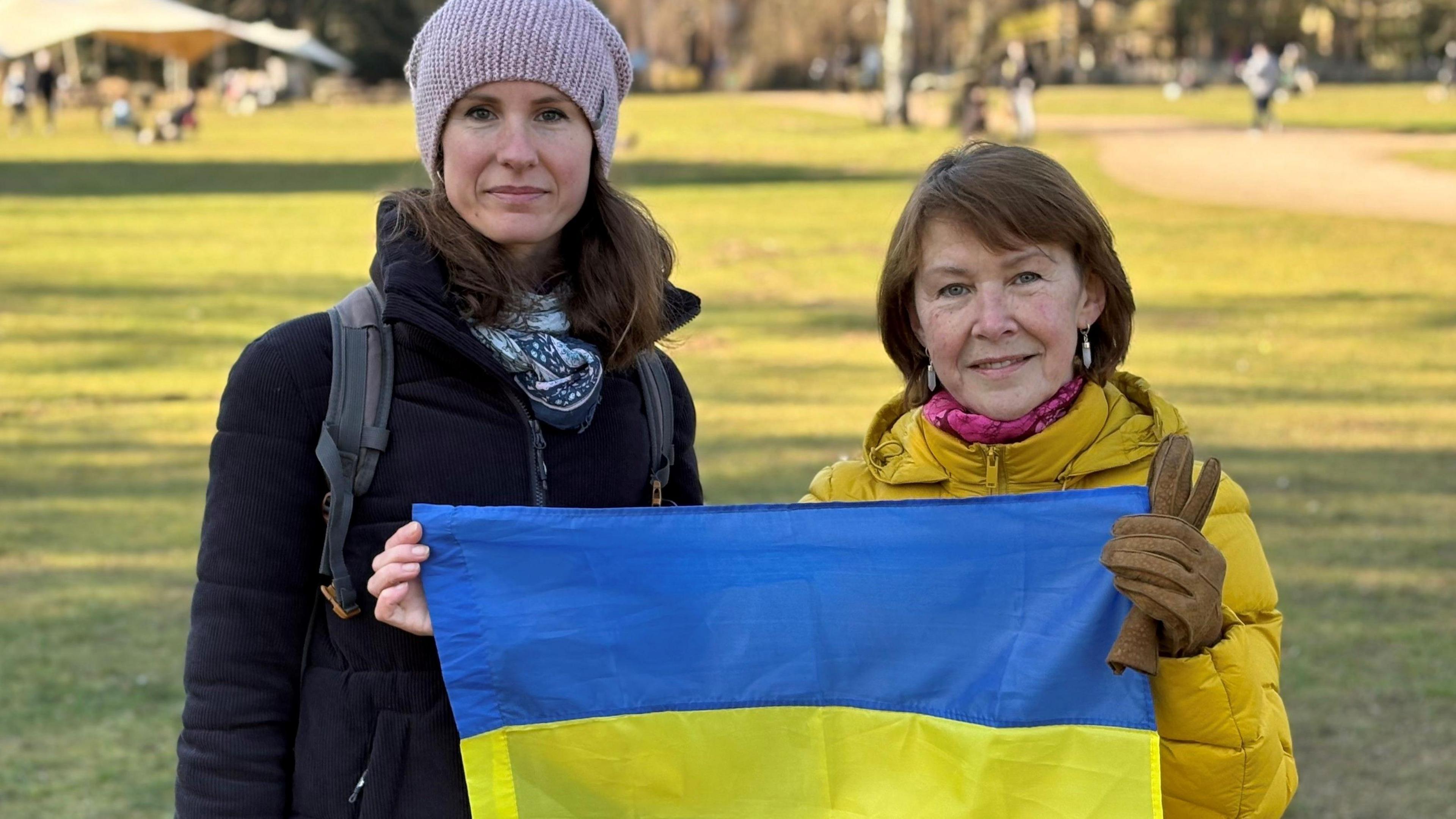 Woman wearing a woolly hat stand next to another in a warm yellow jacket who is also holding a Ukrainian flag. The are on parkland belonging to the Royal Sandringham Estate.