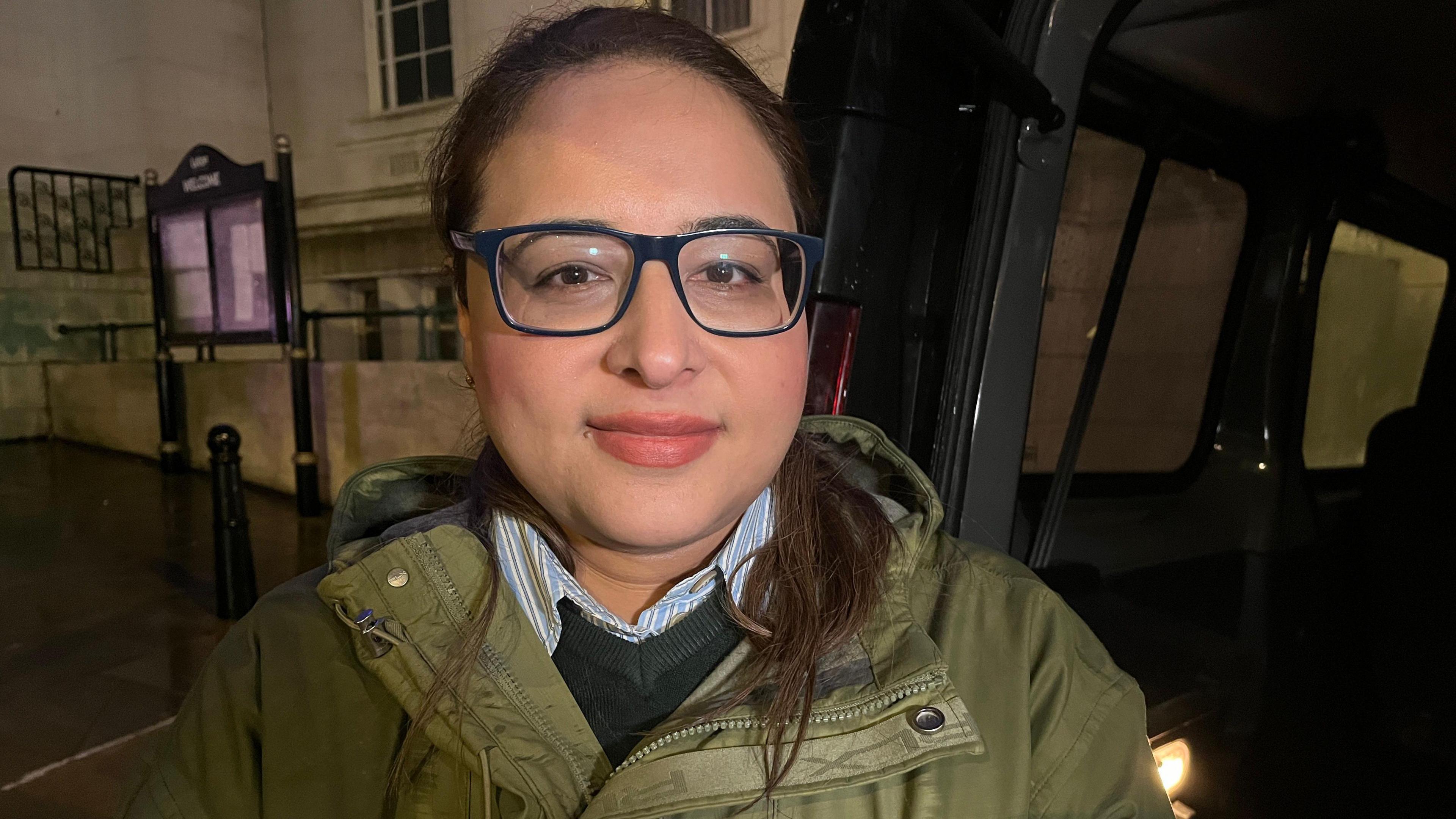 Councillor Javeria Hussai smiles at the camera as she stands outside Luton Town hall after dark. She wears dark blue glasses and has brown eyes and dark brown hair tied back in a pony tail. She is wearing an army green coat, a dark green v neck jumper, and a shirt with blue and white stripes. 