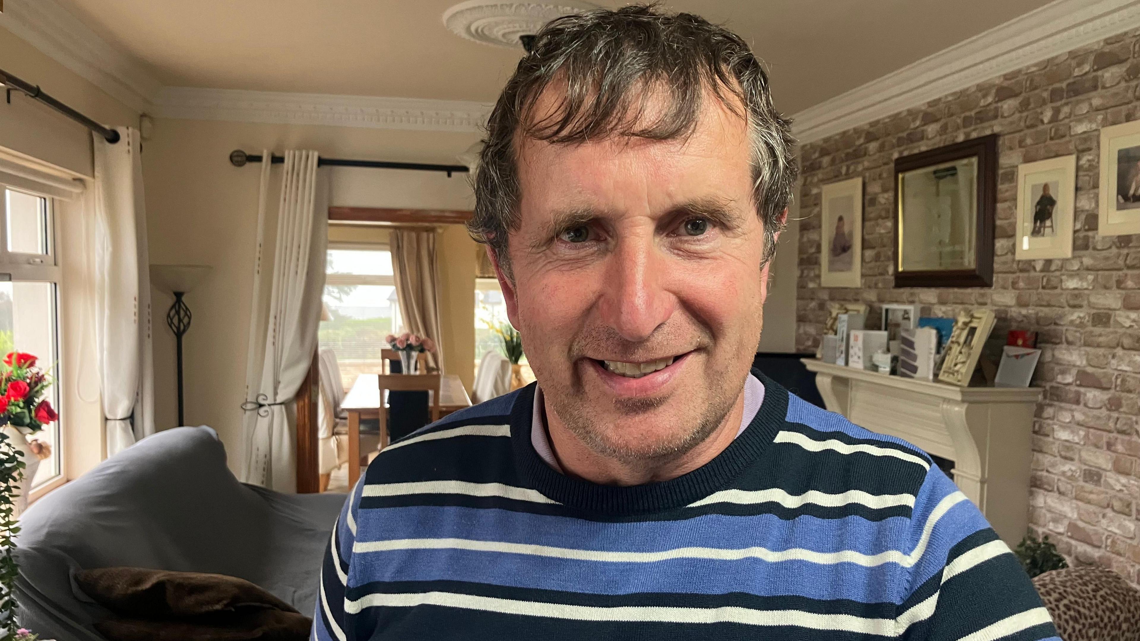 Thomas is standing in a living room. He's wearing a blue, white and black stripped top. There is a mirror hanging above the fireplace. Several frames photos are hanging on the wall and on the fire place. A table is behind him and a sofa.