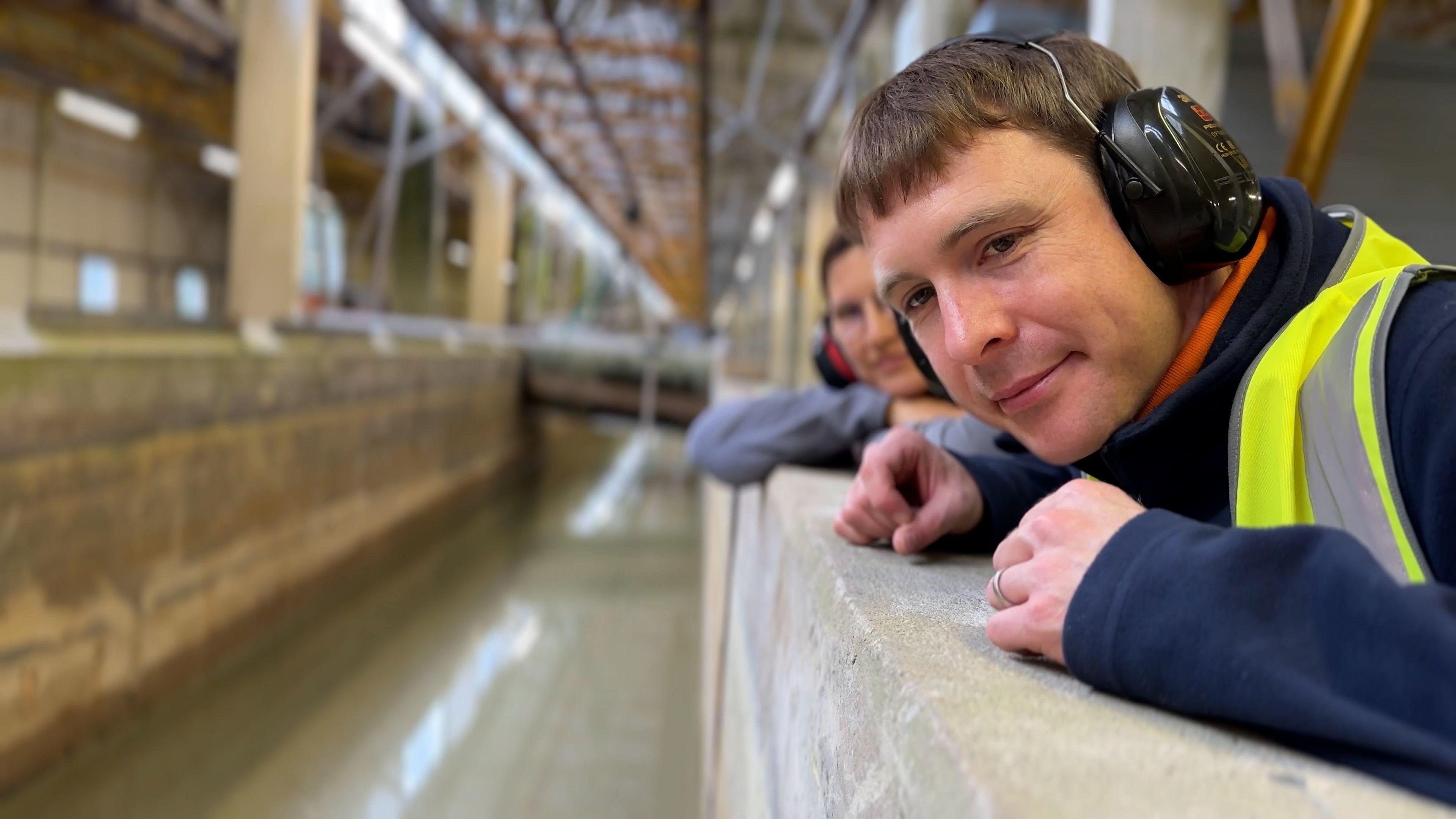 Dr Ian Chandler - a blonde man in a blue fleece and a high-vis jacket leaning over a concrete wall and looking down into a trough of water. There is a woman in soft focus behind him, also looking at the water, and they appear to be in a large building similar to an aircraft hangar. Both people are wearing ear defenders.