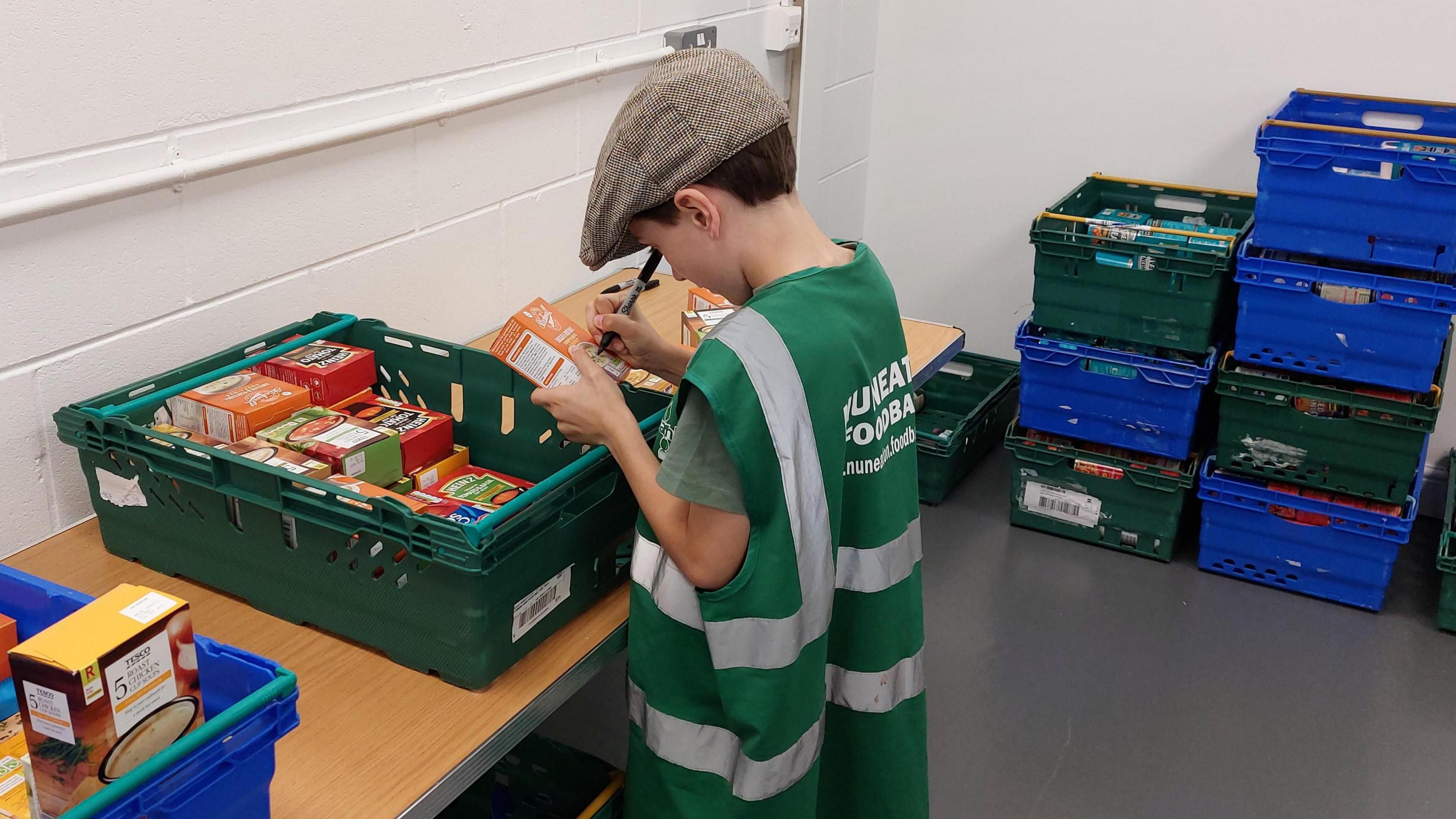 A child wearing a flat cap, a green t-shirt and an adult-sized work vest with Nuneaton Foodbank branding is labelling products from a green plastic crate. There are blue and green plastic crates stacked up in the room to the side of the child.