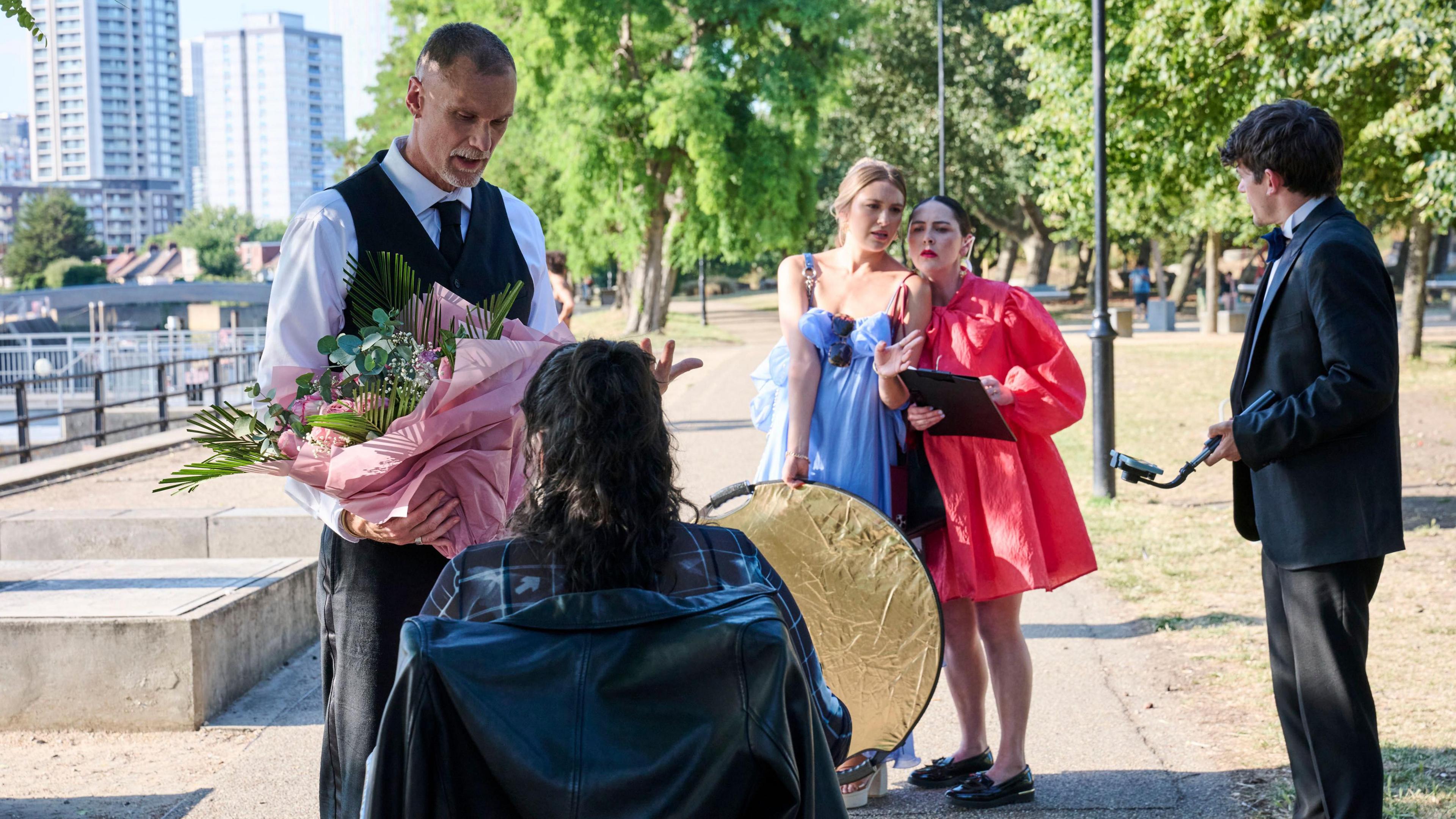 In a park, a man in a suit gives flowers to a woman using a wheelchair who is wearing a leather jacket. On their right a woman in a blue dress is holding a lighting prop and a woman in a red dress is holding a clip board. Next to them is another man in a suit holding a selfie stick.