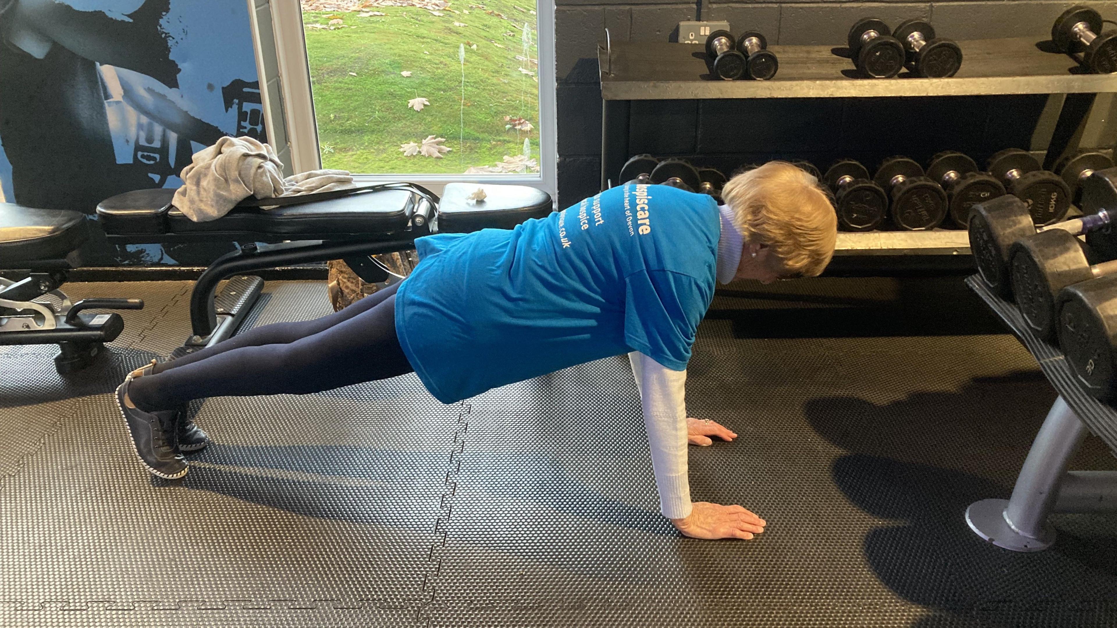Dorrie is doing a plank with perfect form. She is wearing navy leggings and a blue T-shirt. There are racks of dumbbells of different sizes in the background.
