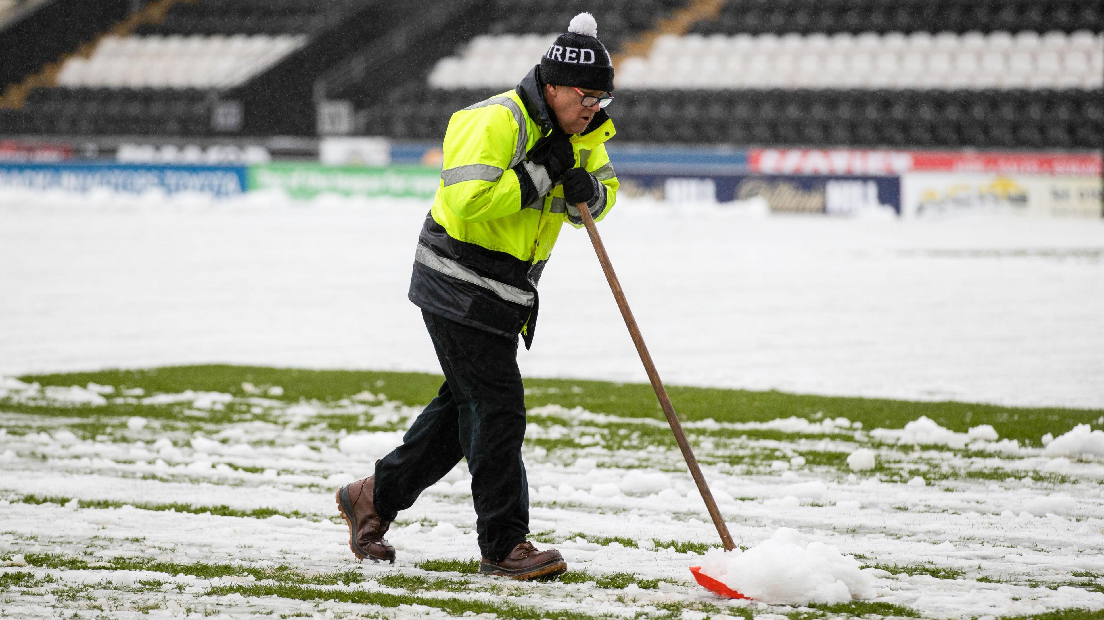 A man wearing a hi-vis jacket and a bobble hat, clears snow off a football pitch with a shovel