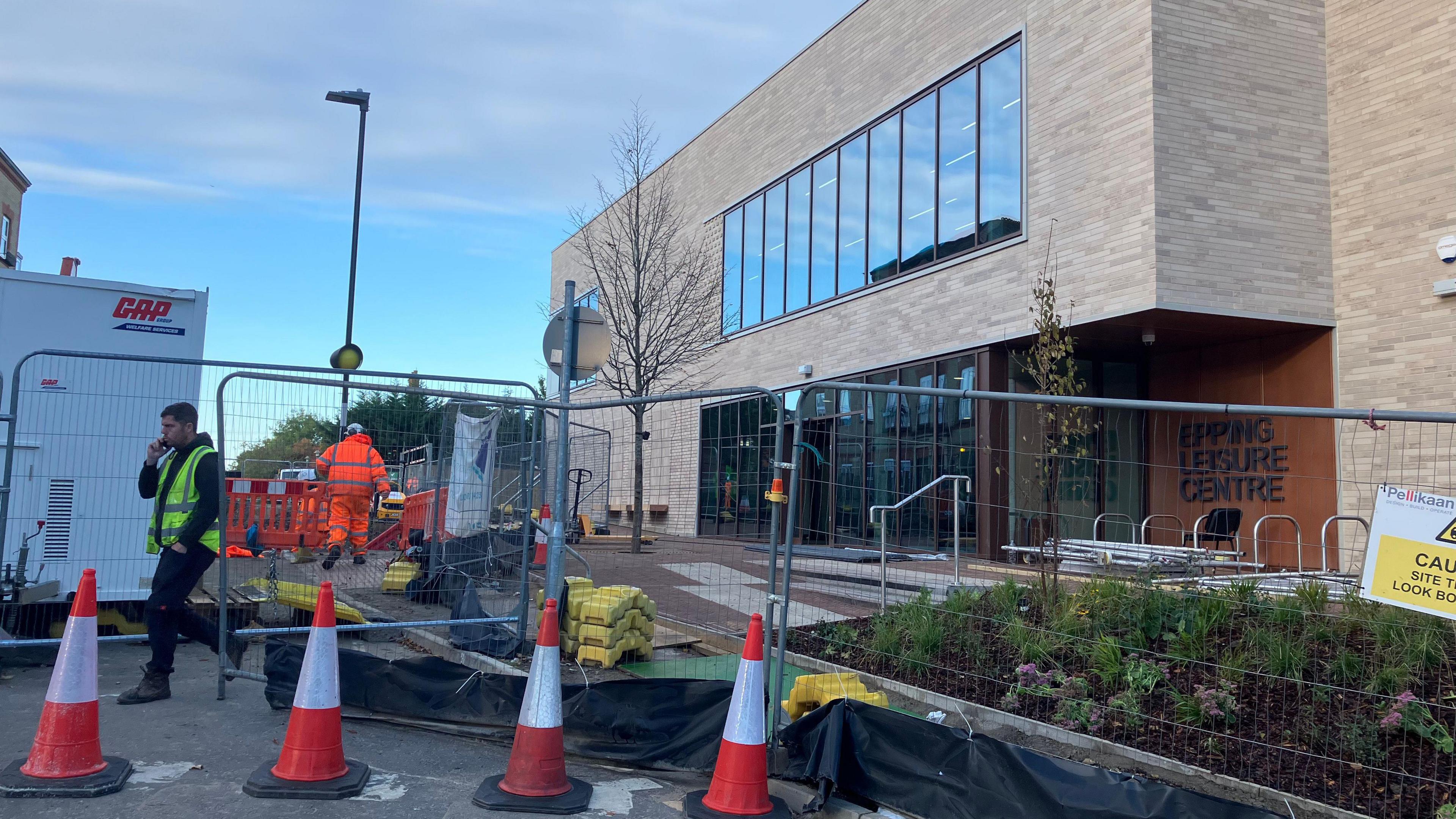Some red and white cones in front of a metal fence that's cordoning off a large leisure centre. A man in a high vis jacket is walking across the shot