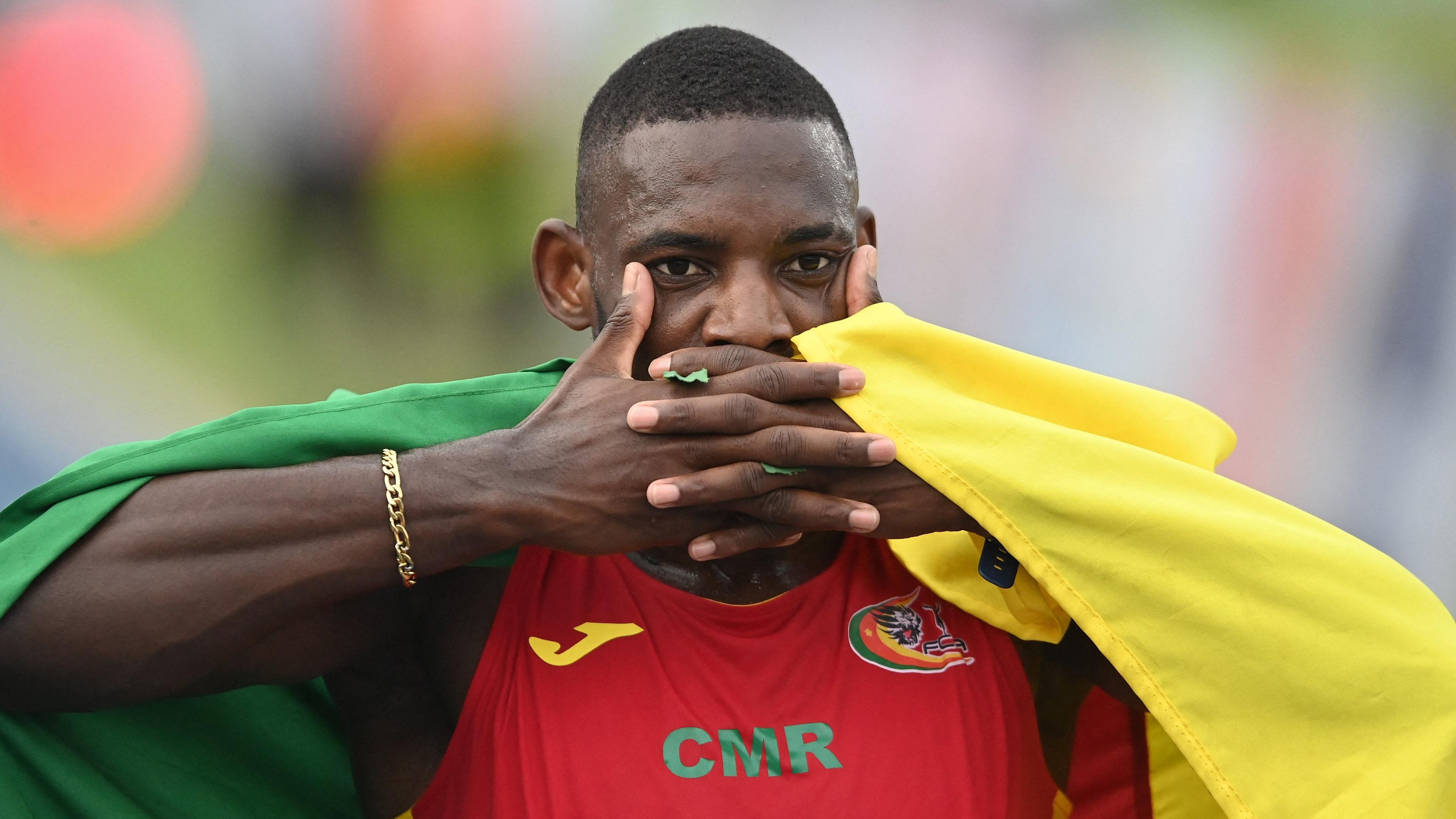 Emmanuel Eseme, seen from chest up wearing a red vest with the letter CMR and a Cameroon crest on it, holds a Cameroon flag draped over his back and covers his mouth with interlocking fingers to mimic the goal celebration of Arsenal striker Viktor Gyokeres