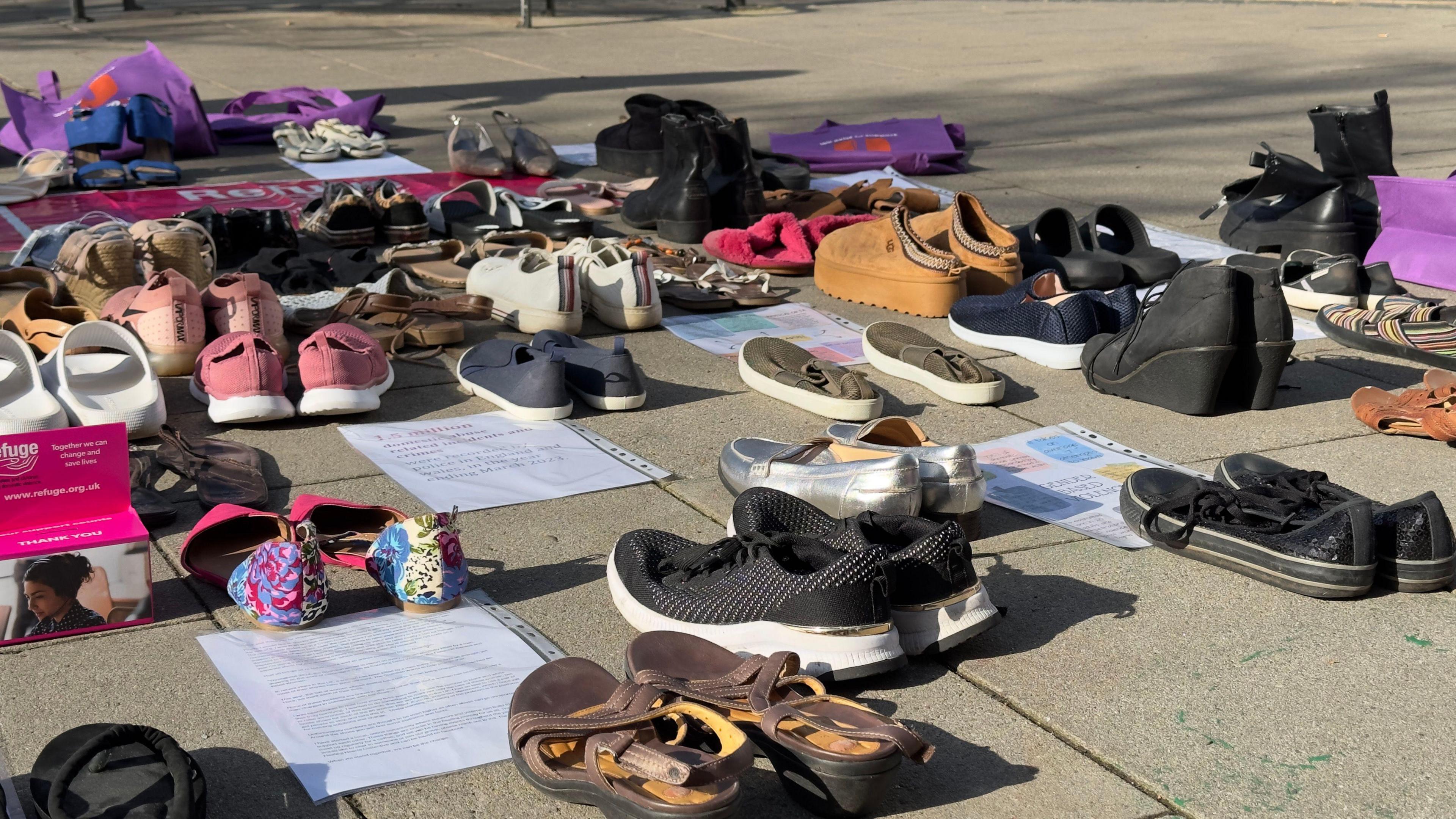 Dozens of pairs of shoes are placed in rows, some alongside printed literature in plastic sleeves, on a pavement.