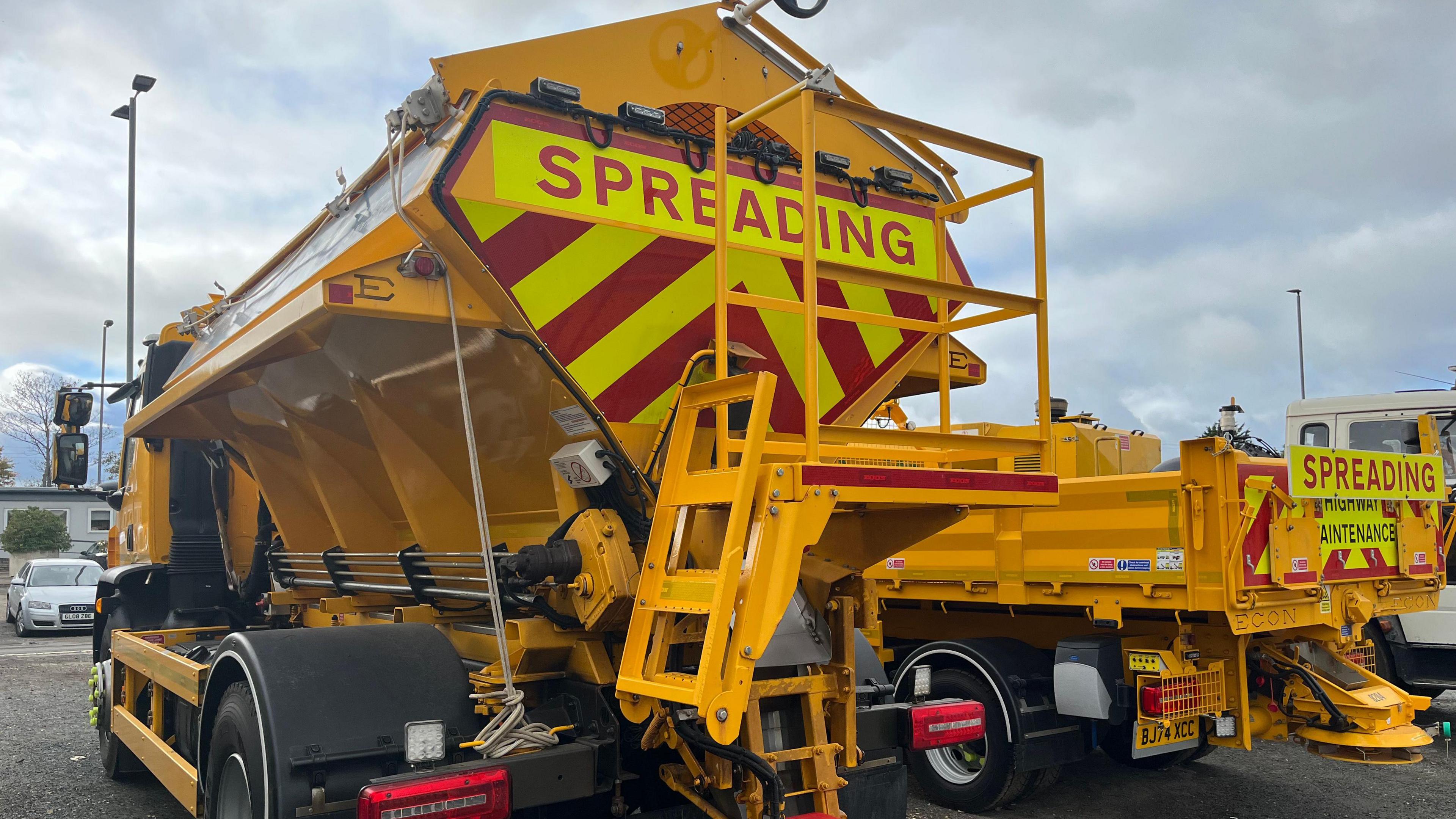 The back of a yellow gritting lorry parked in a car park. It has a sign saying SPREADING and yellow and red stripes across the back of its tanker.