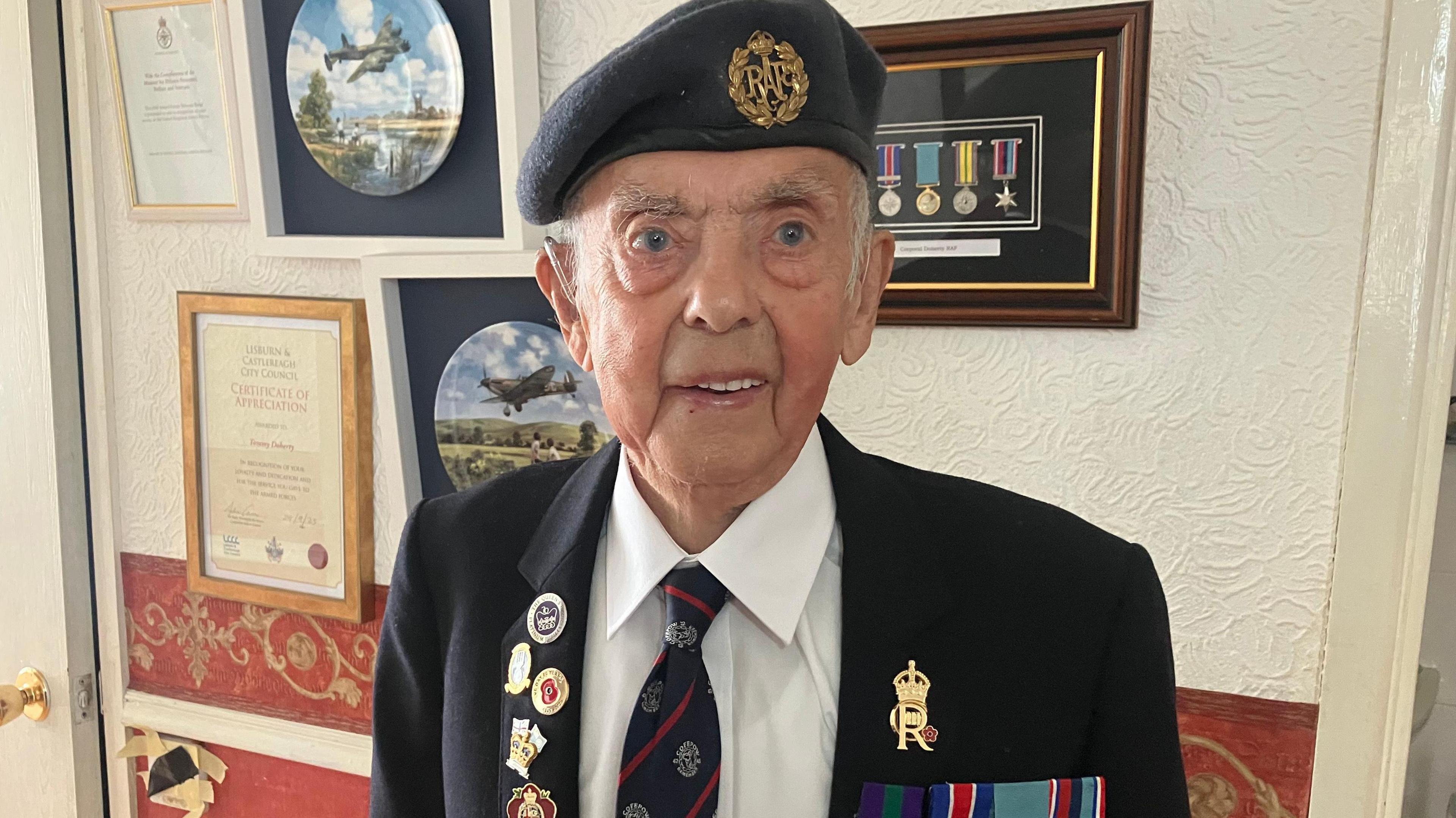 Tommy Doherty  stands in a room in his home, in front of a wall on which medals and a number of plates displaying images of the war are scene. Tommy is wearing a war blazer and beret, and a shirt and tie. He wears a number of military badges and medals on his blazer.