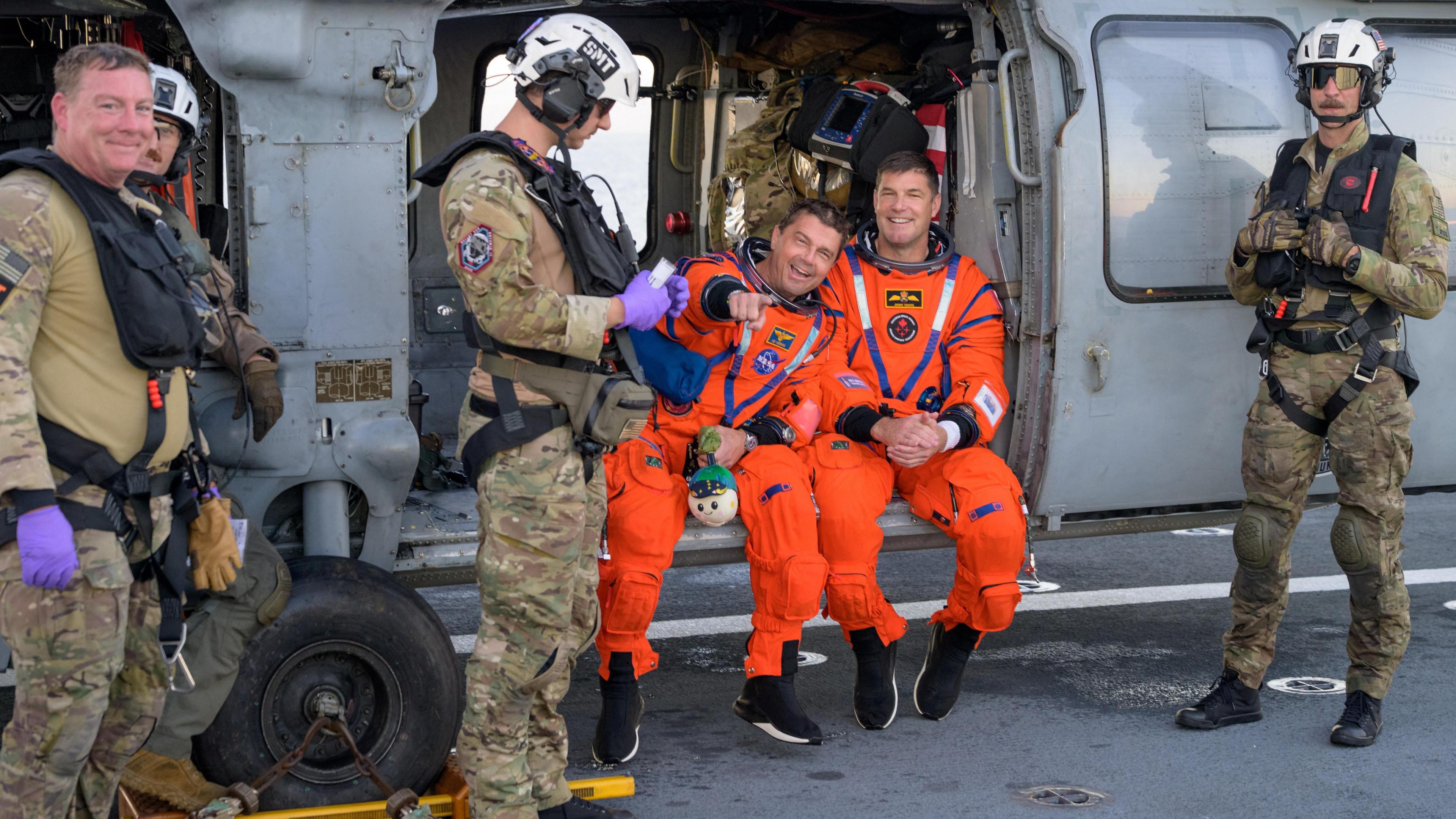 Astronauts Reid Wiseman and Jeremy Hansen sit smiling in open helicopter in their orange suits with helicopter crew around them. 