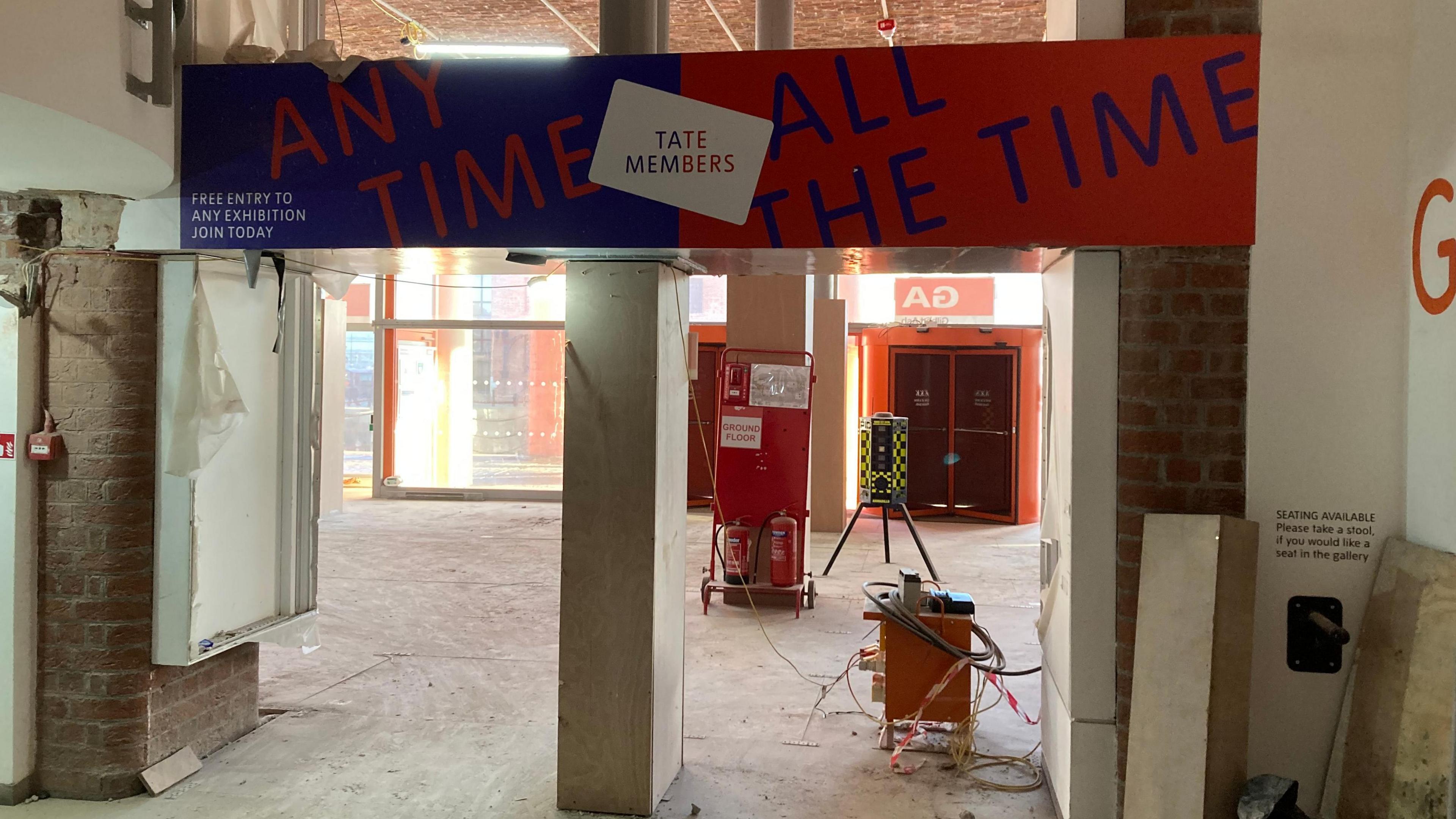 A red and blue sign that reads 'Tate members, any time all the time' hangs over an empty doorway. The floor and brick is exposed on the wall.
