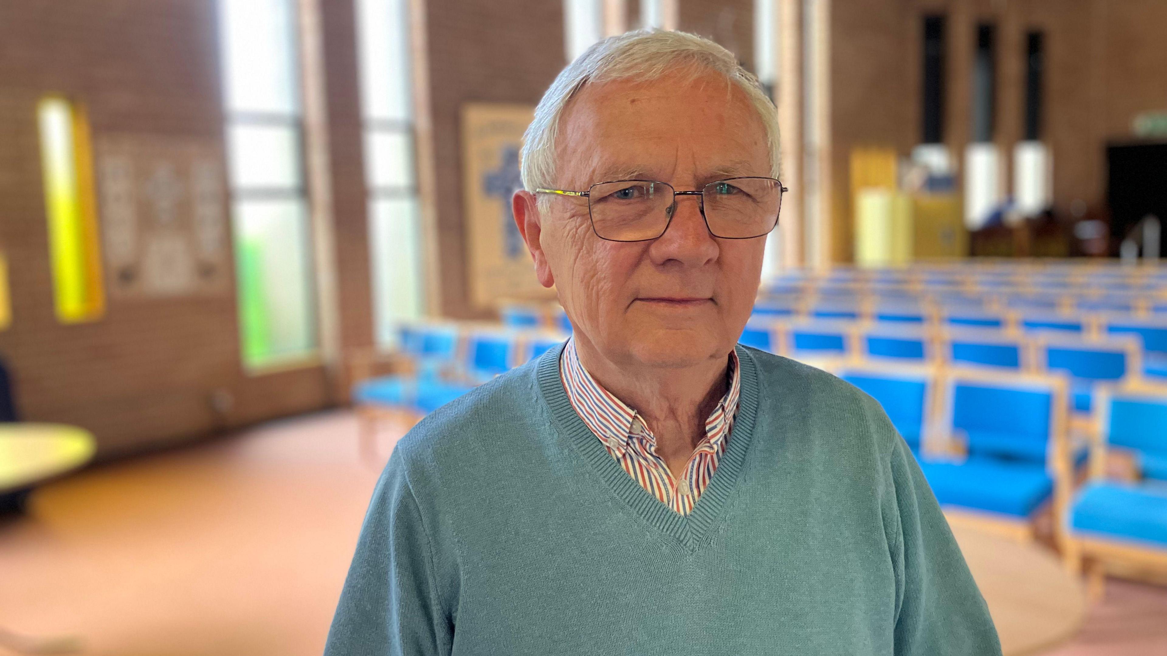 Philip wearing blue v-neck jumper and a checked shirt inside - standing inside the main prayer hall of the church- with blue chairs in the background