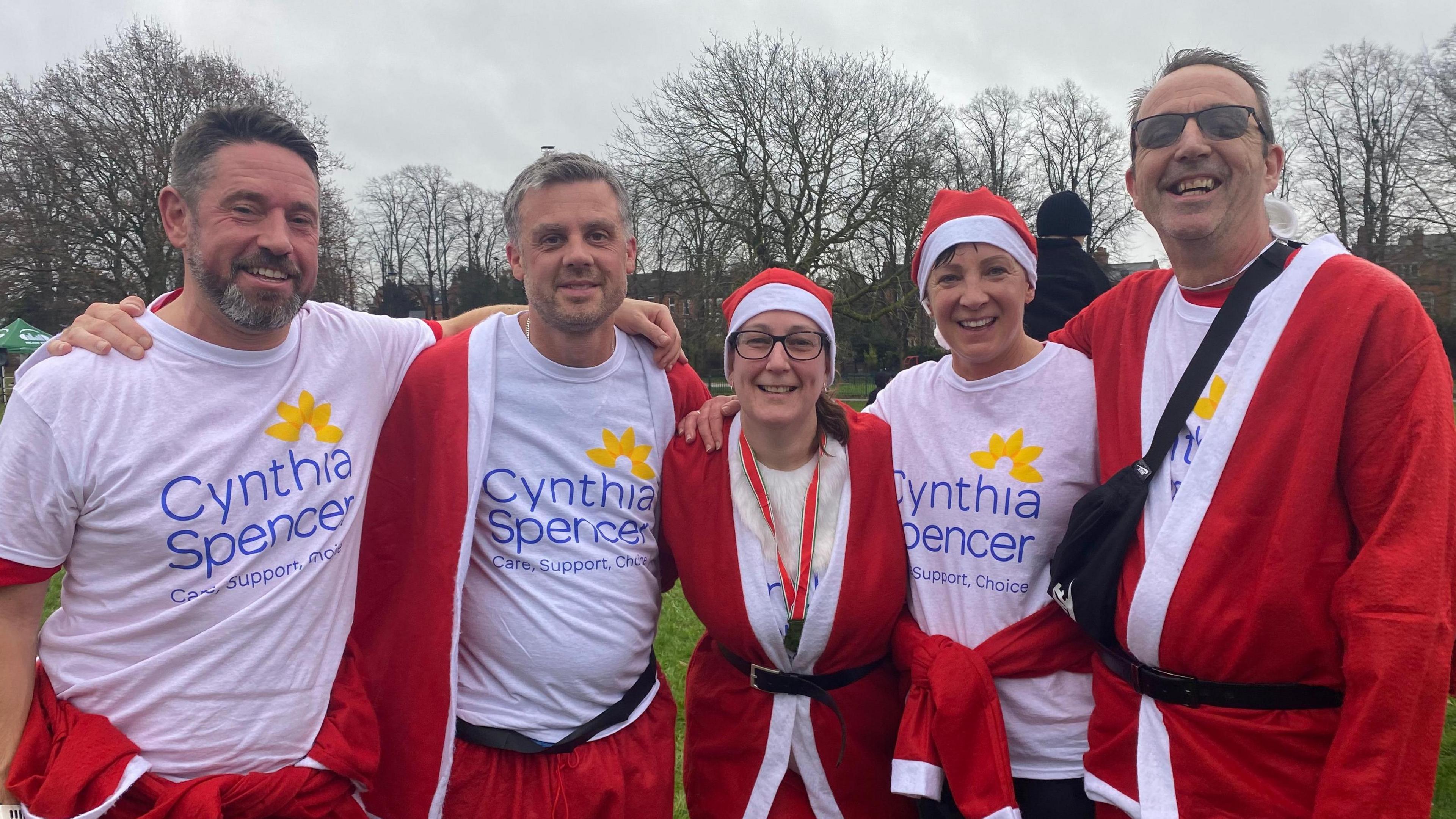 A group of five people in a line staring at the camera. They are dressed as Santa with each of them wearing a white top with the words ' Cynthia Spencer' underneath the red and white jacket.