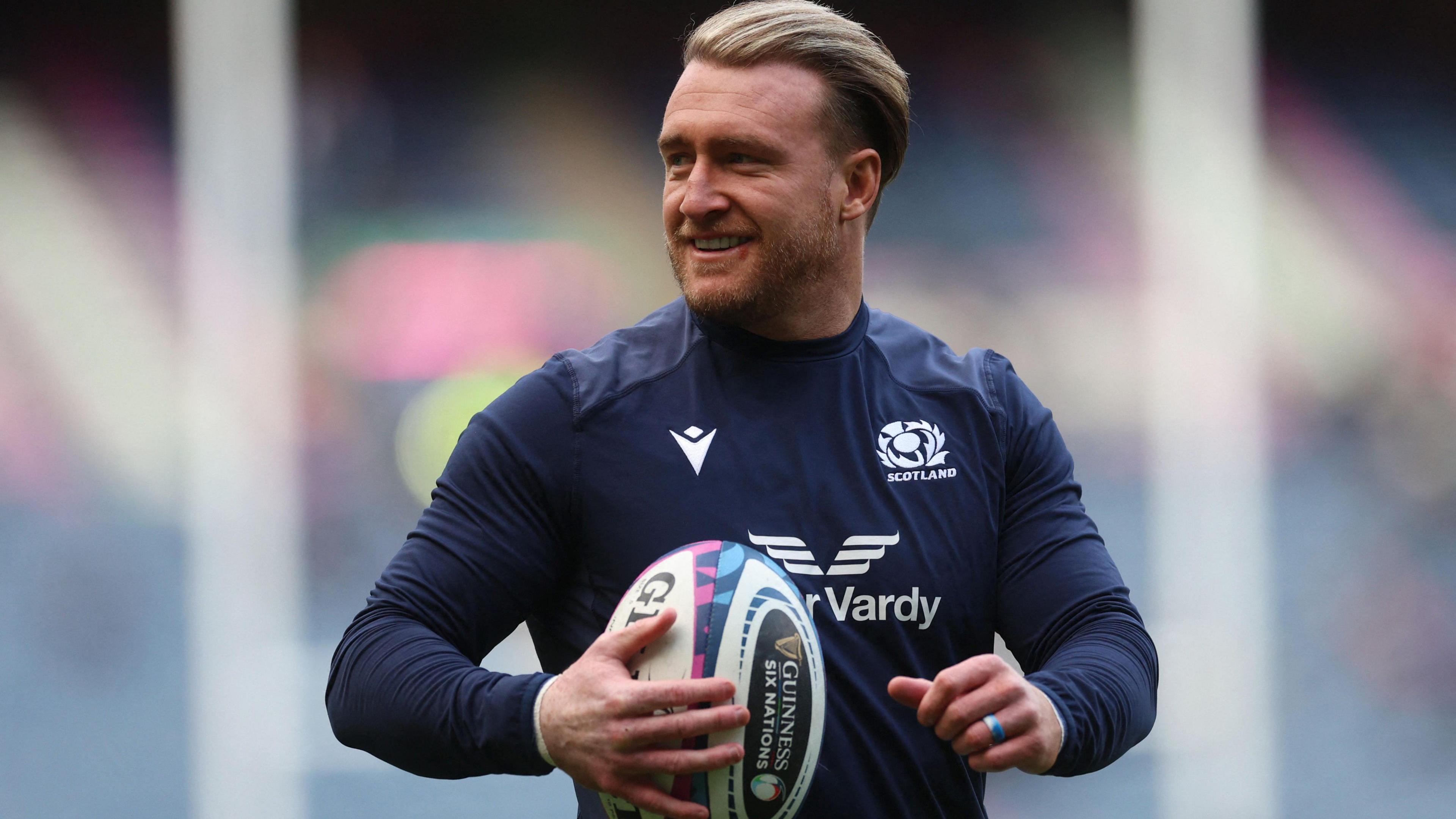 Stuart Hogg wearing a blue Scotland kit holding a rugby ball. He has blonde hair which is brushed back.