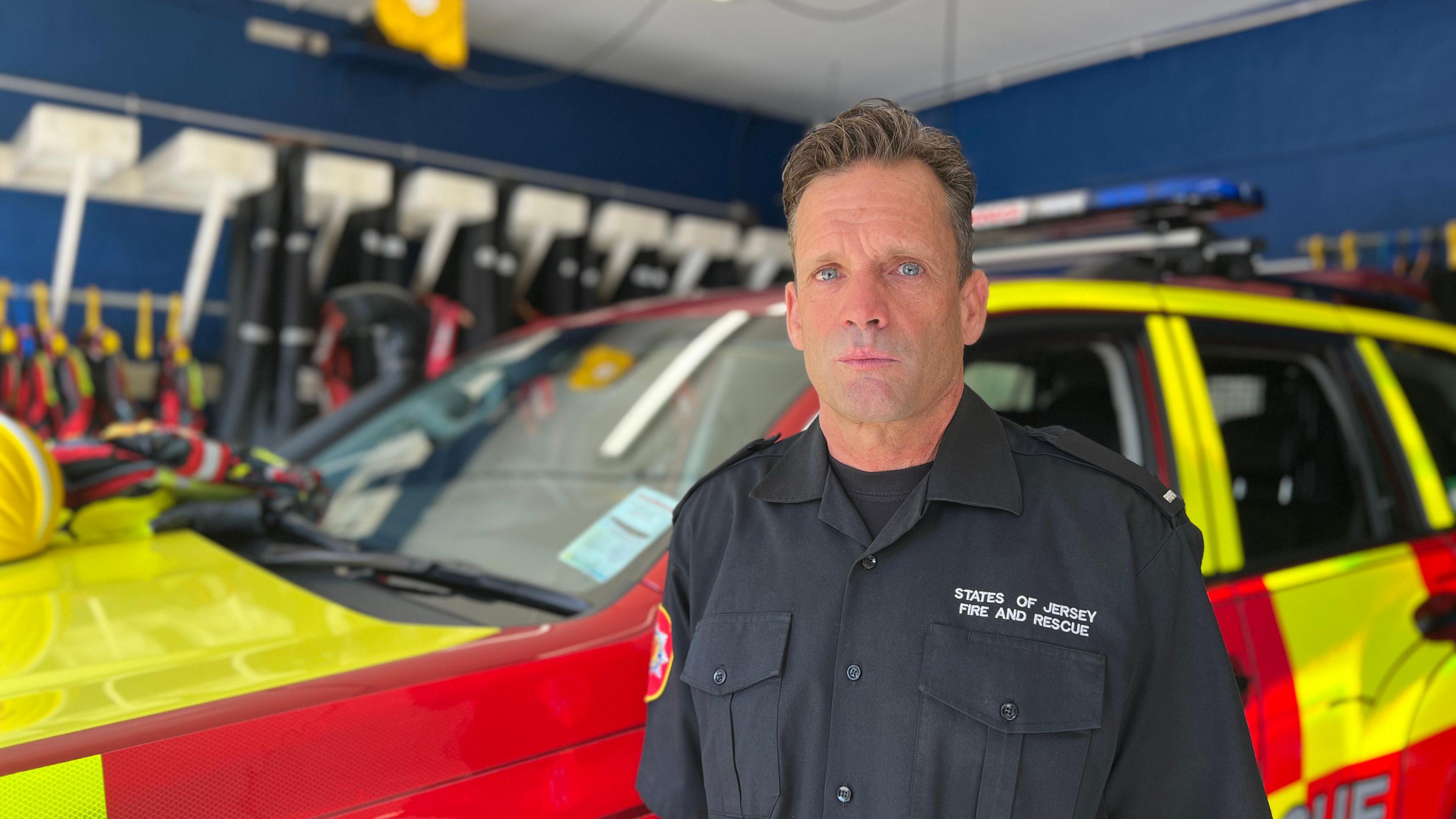 Nick looks at the camera as he stands in front of the yellow and red 4x4 rescue vehicle. He is wearing a States of Jersey Fire and Rescue shirt with a black T-shirt underneath it. Behind the vehicle, sea rescue swimming gear is hanging in the background.