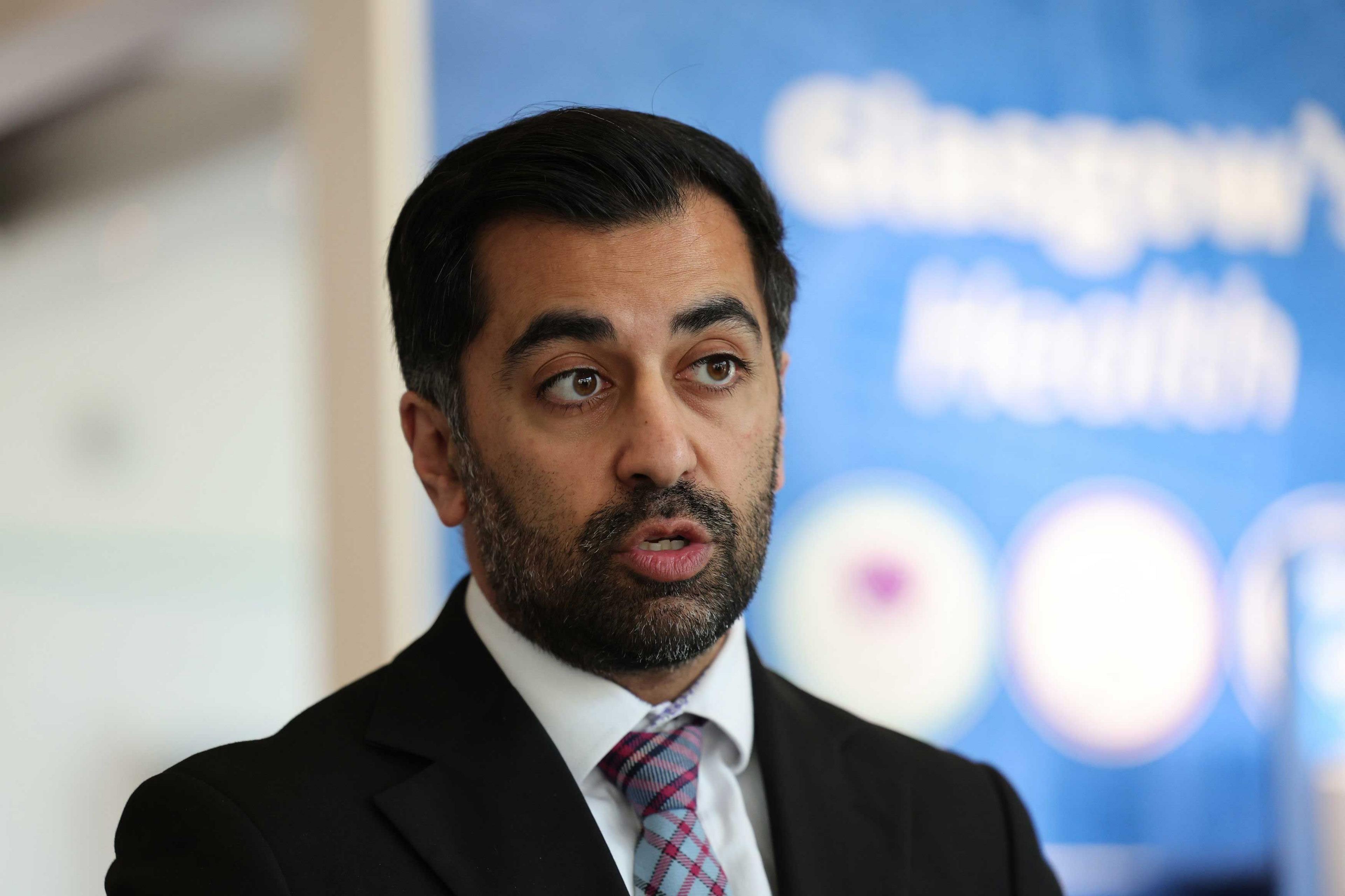 Humza Yousaf making a speech, wearing a black suit, white suit and a tartan tie. There is a blue background, which has been blurred.