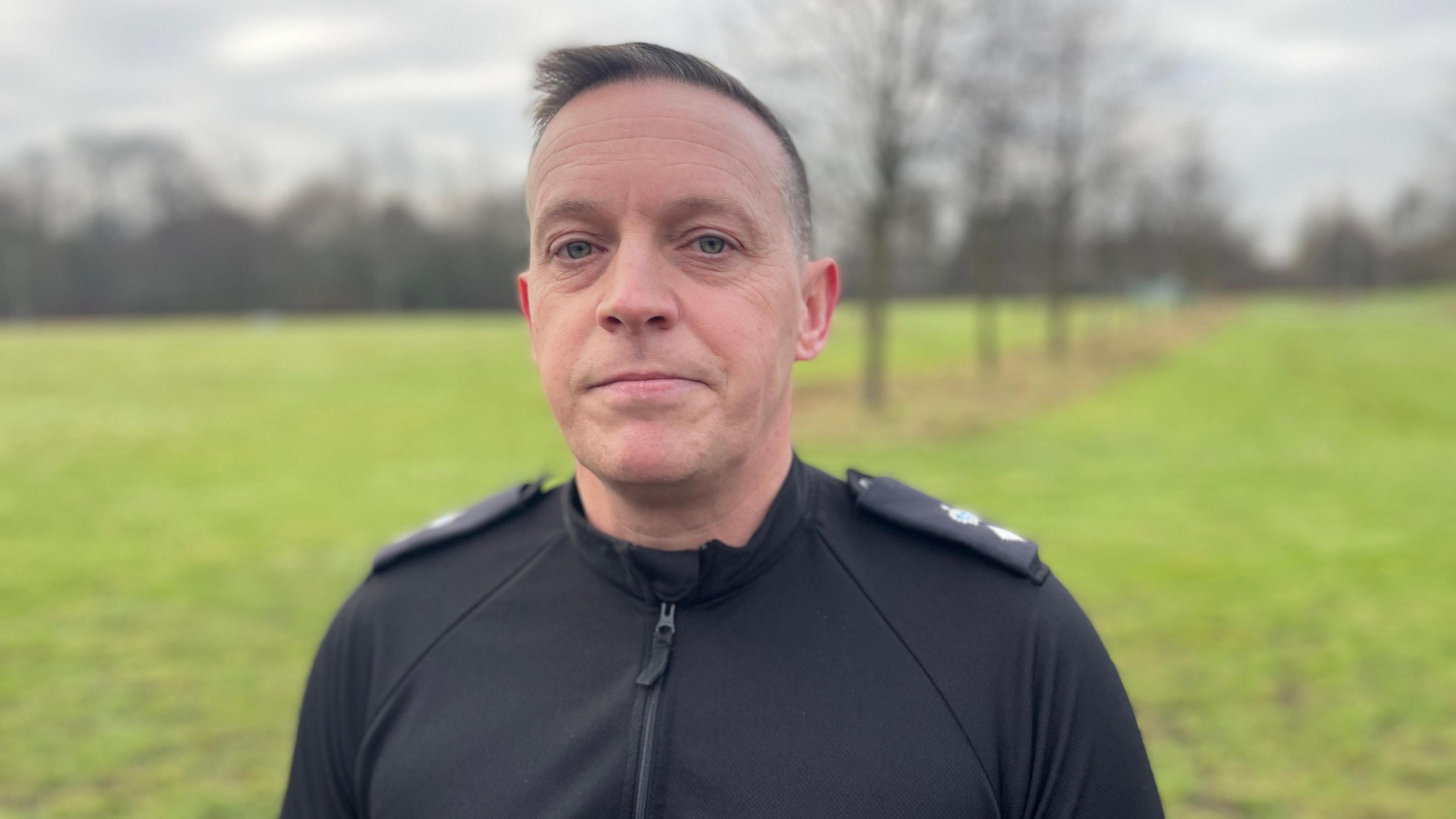 Ch Supt Gregg Banks looking right at the camera. He has short, dark hair and is wearing a black top. He is standing in front of a blurred background of trees and grass.