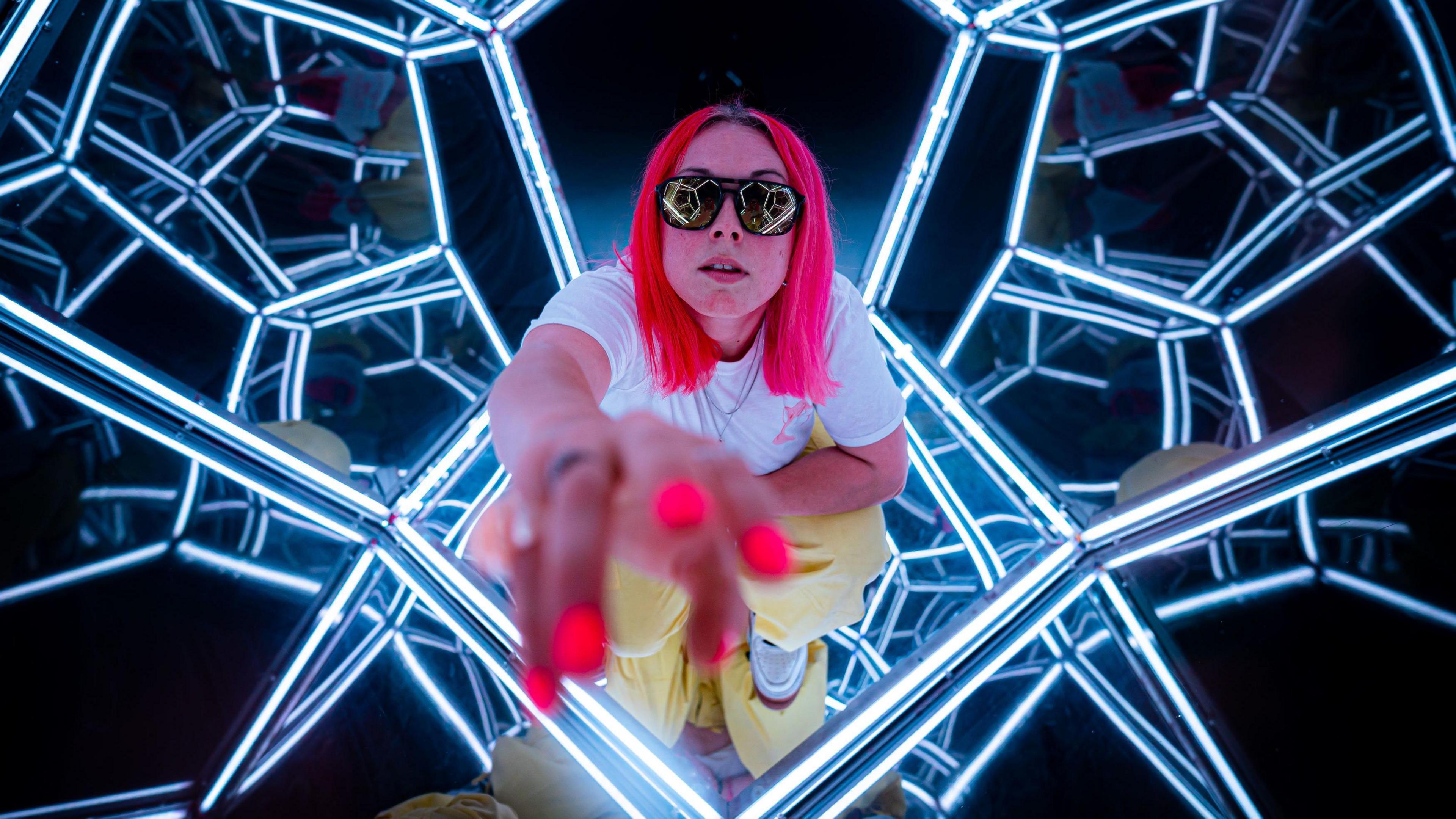 A woman with pink hair, pink sunglasses and pink nails plays around with her reflection inside a mirrored structure 