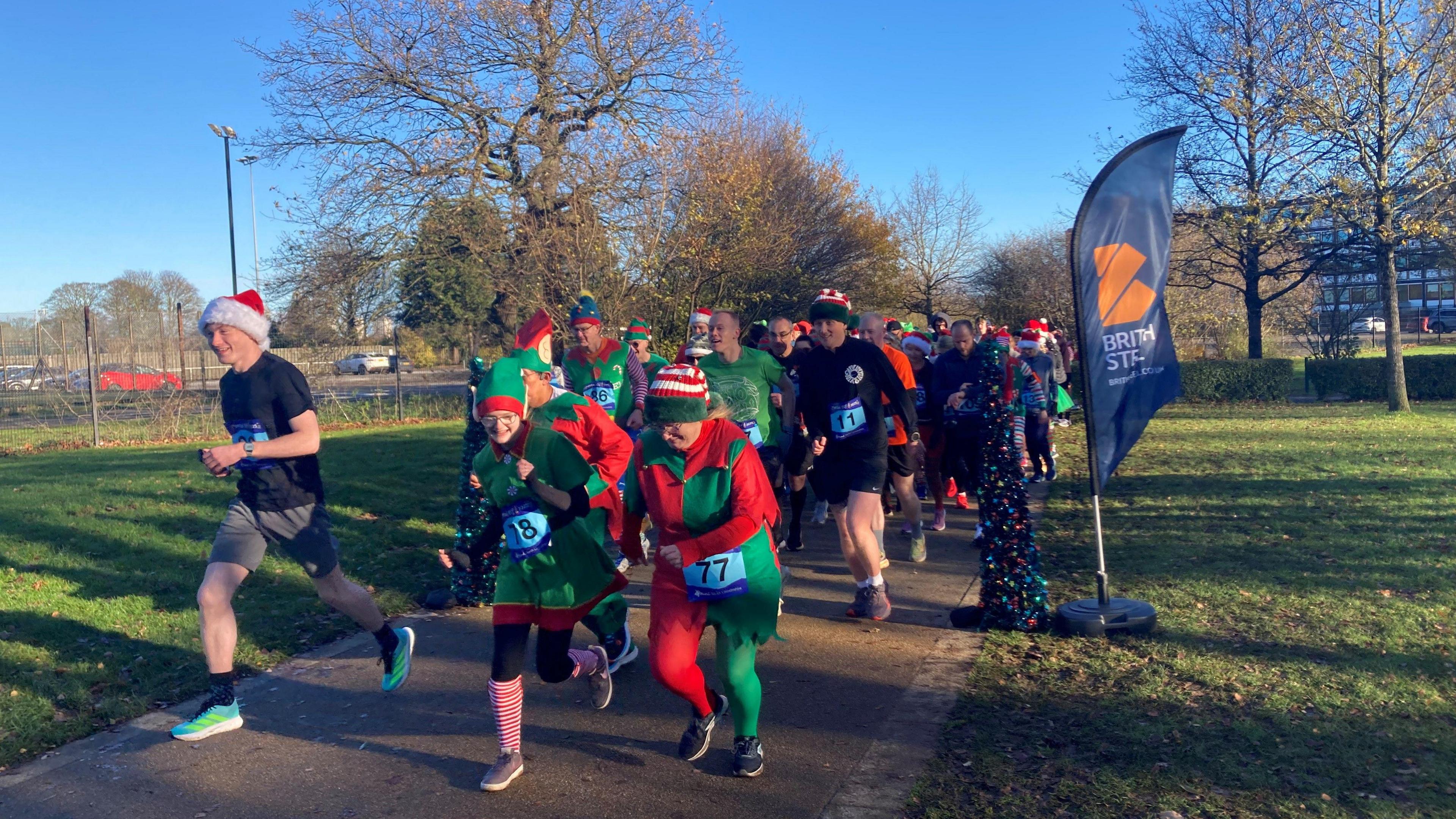 Runners dressed in elf outfits are running through the park. The sky is bright blue and the sun high lights the colours well.