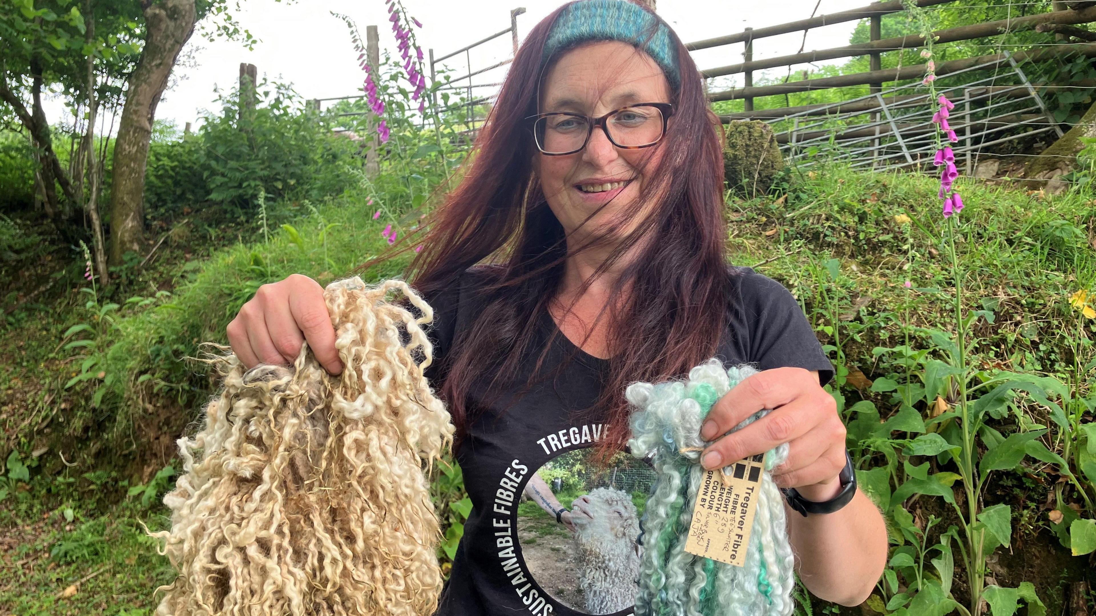 A woman with long brown hair and a woven mohair band holds up curly sheep wool that is undyed and compared with her own dyed fibre.