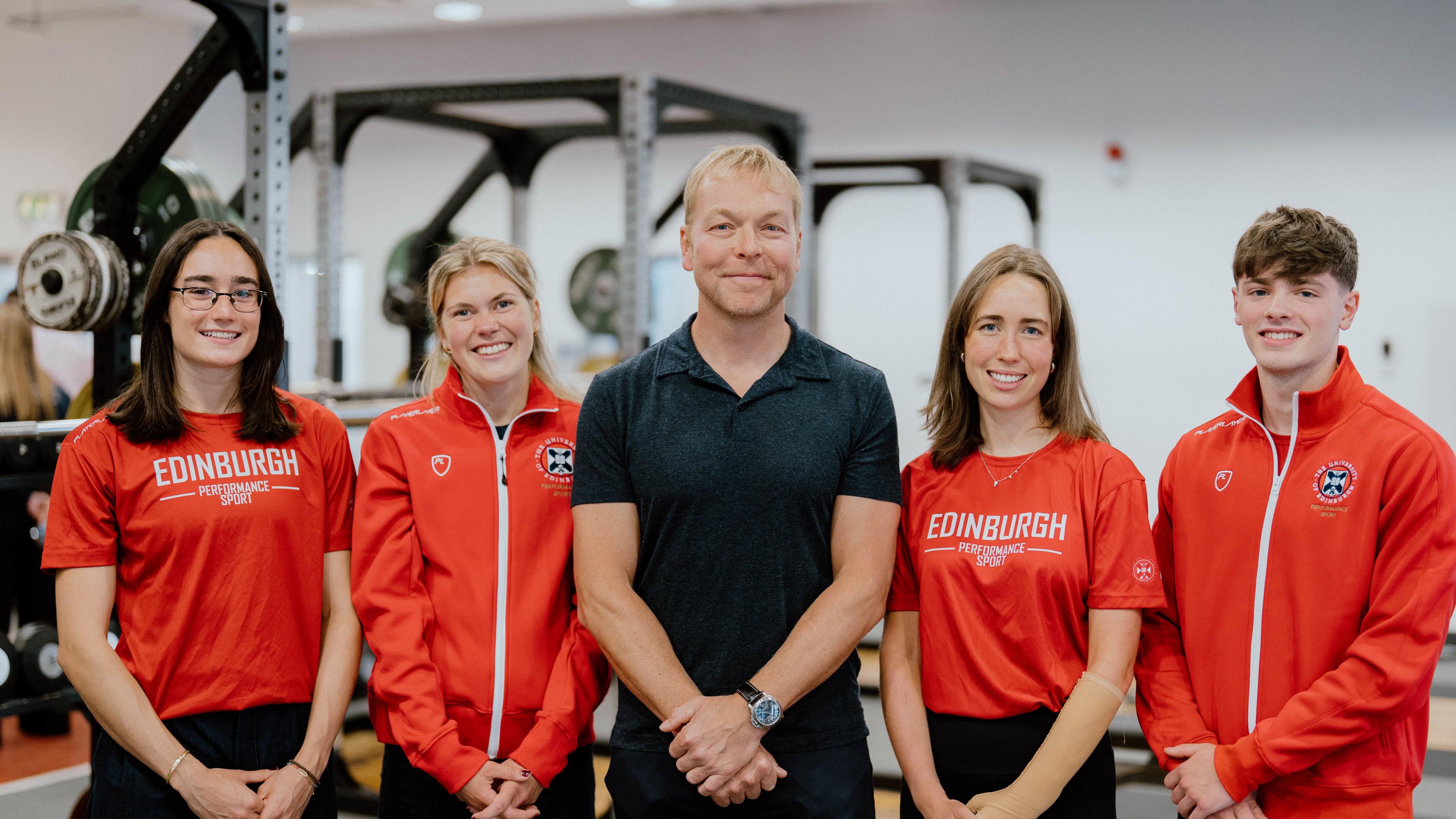 Three young women and a young man stand on either side of Sir Chris Hoy. Sir Chris is dressed in a black polo shirt and trousers. The students are wearing red Edinburgh University t-shirts and tracksuits. 