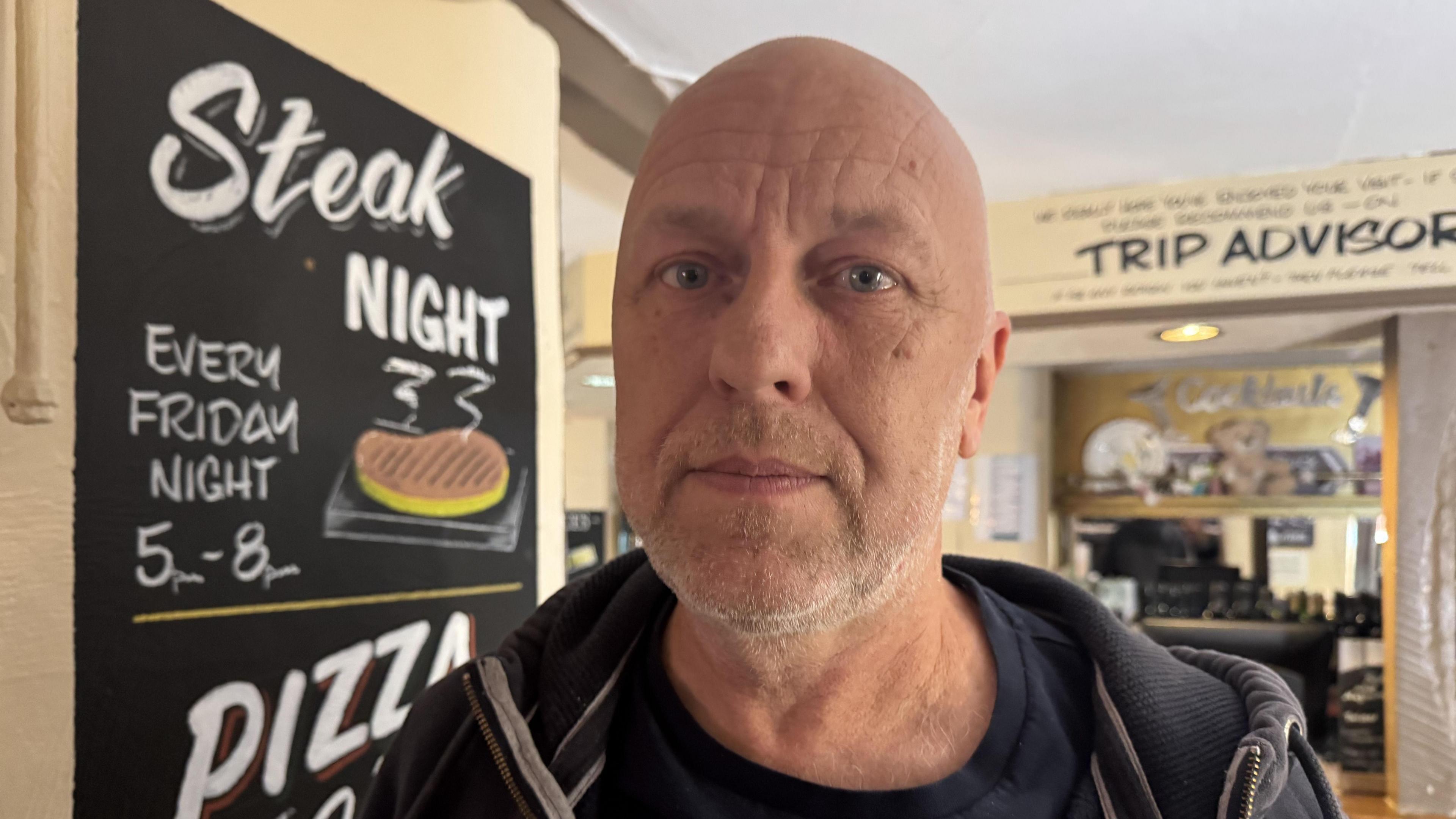 A man wearing a hoodie stood inside a pub. There are signs on the wall behind him and writing on another next to him.